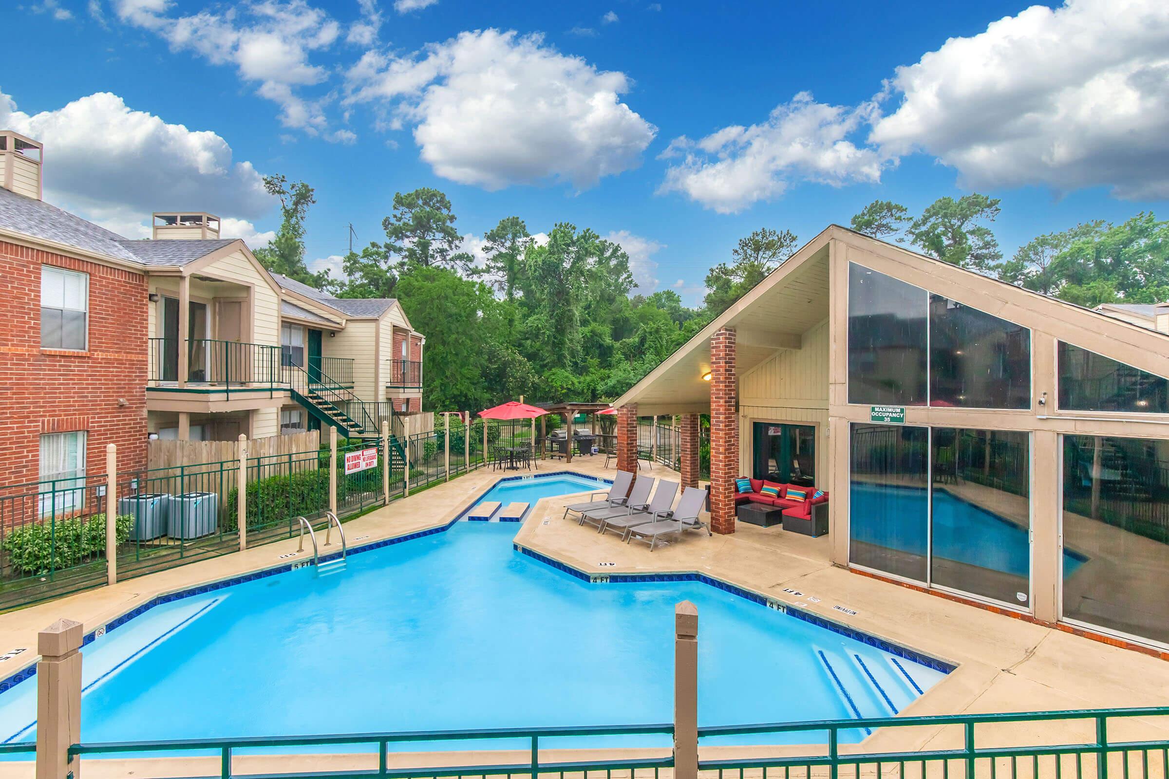 A view of a residential swimming pool area with a clear blue pool, surrounded by lounge chairs and shaded by umbrellas. Nearby, there are two buildings with outdoor staircases and a covered seating area with large windows, set against a backdrop of green trees and a vibrant blue sky with fluffy clouds.