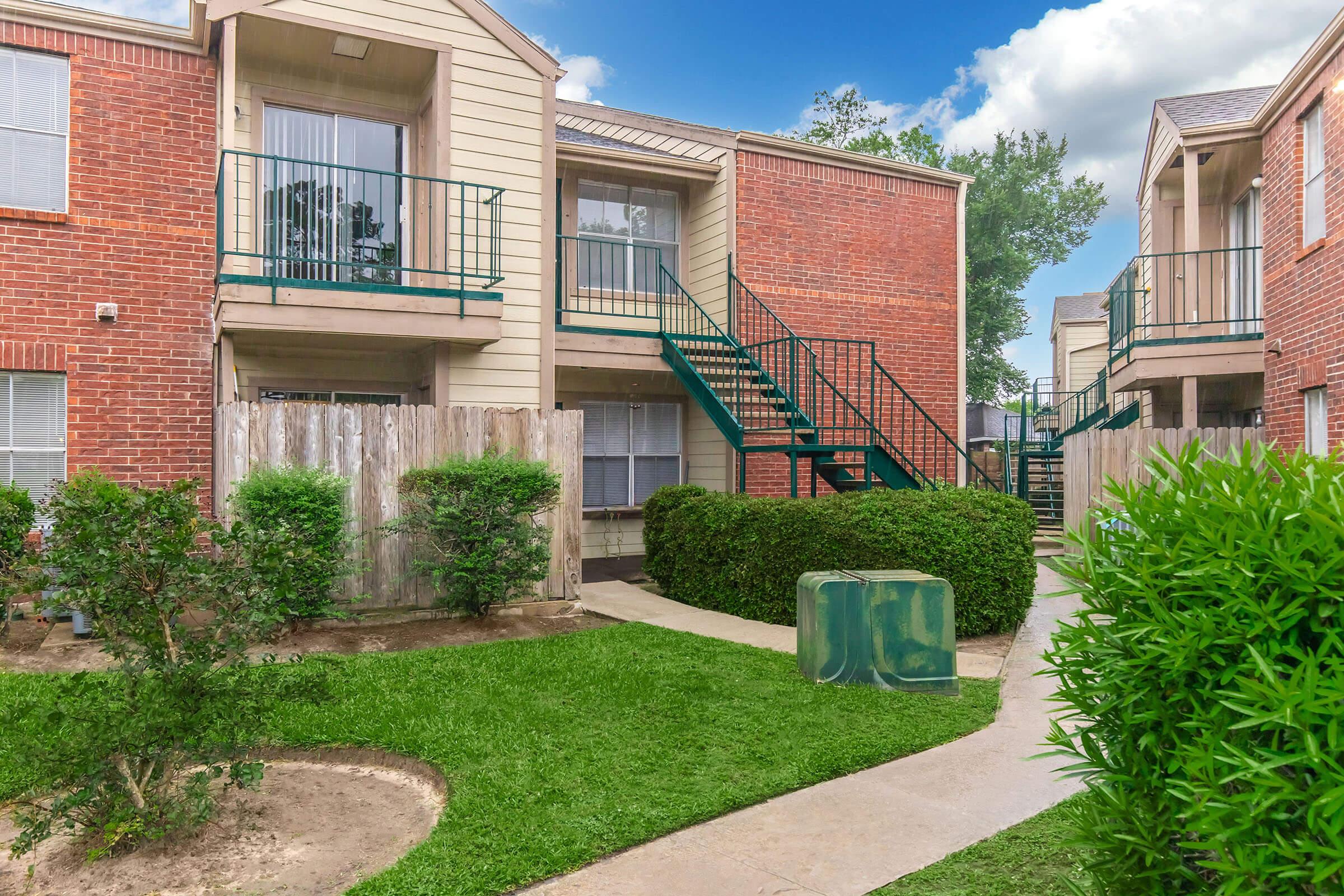 A view of a two-story apartment complex with brick exteriors and green staircases. The courtyard features manicured grass, small shrubs, and a pathway leading between the buildings. There is a wooden fence and some landscaping elements, creating a peaceful residential environment.
