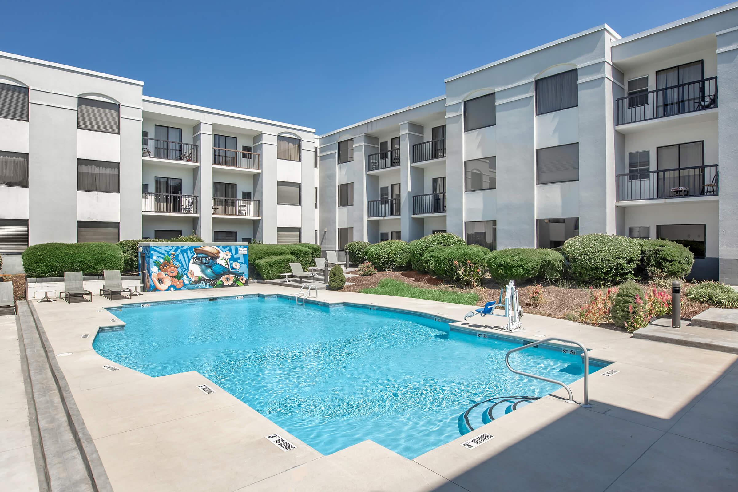 A bright and inviting apartment complex featuring a swimming pool surrounded by well-manicured greenery. The pool area includes lounge chairs and a decorative mural on the wall of the adjacent building. There are three-story apartment buildings in the background under a clear blue sky.