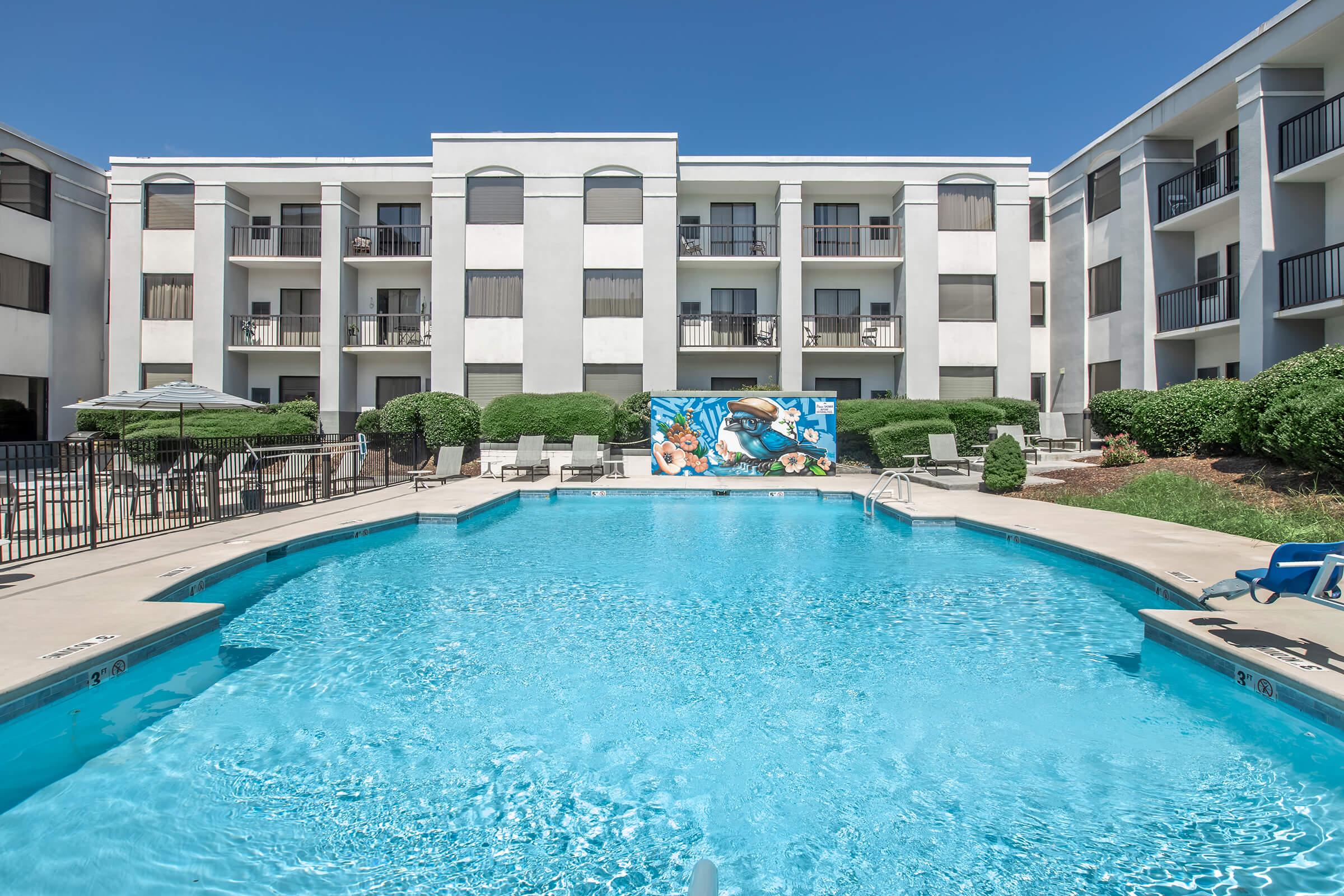 A clear swimming pool surrounded by lounge chairs and shaded seating areas, with a colorful mural on the building in the background. The building features multiple balconies and large windows, set against a bright blue sky. Lush greenery adds to the inviting atmosphere.