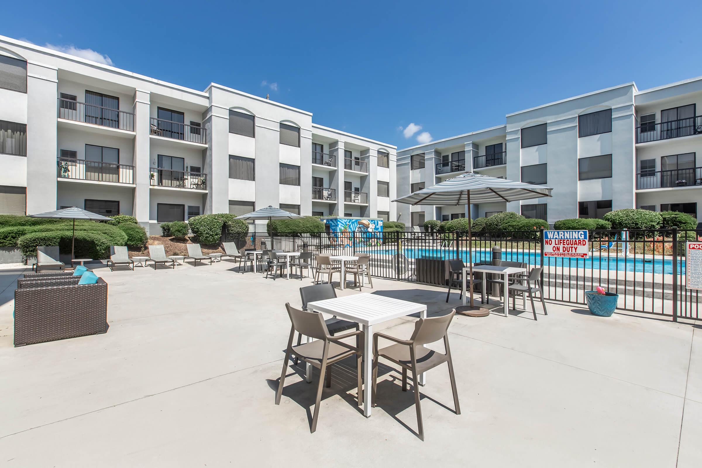 A bright outdoor space featuring a swimming pool surrounded by lounge chairs and tables. The area includes a gazebo and is bordered by a modern apartment building with balconies. Lush greenery adds to the ambiance, and a warning sign is visible near the pool. The sky is clear and blue, enhancing the inviting atmosphere.