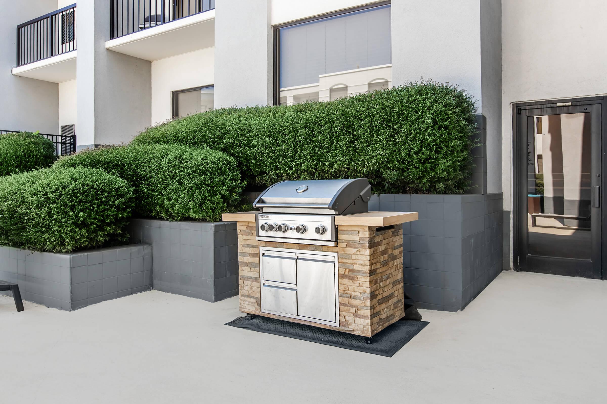 An outdoor grilling station featuring a stainless steel gas grill on a stone base, placed on a patio surrounded by neatly trimmed hedges and a building. The area is well-lit and has a modern, clean look, ideal for barbecues and outdoor cooking.
