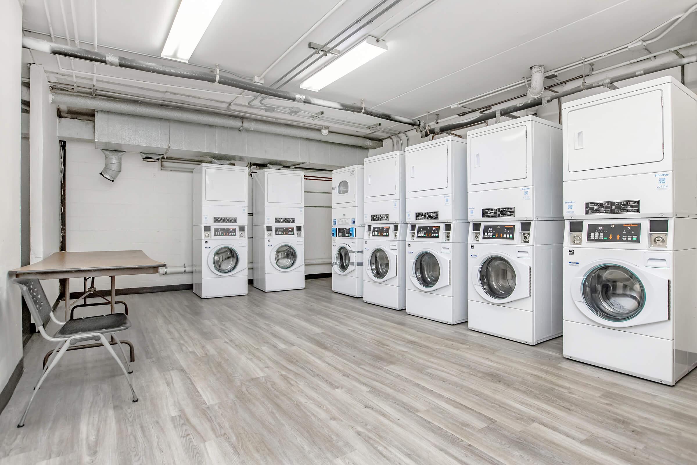 A clean, modern laundry room featuring several stacked washer and dryer units. The space has a smooth, light-colored floor, exposed pipes on the ceiling, and a single chair at a table against the wall, creating a tidy and functional environment for laundry activities.