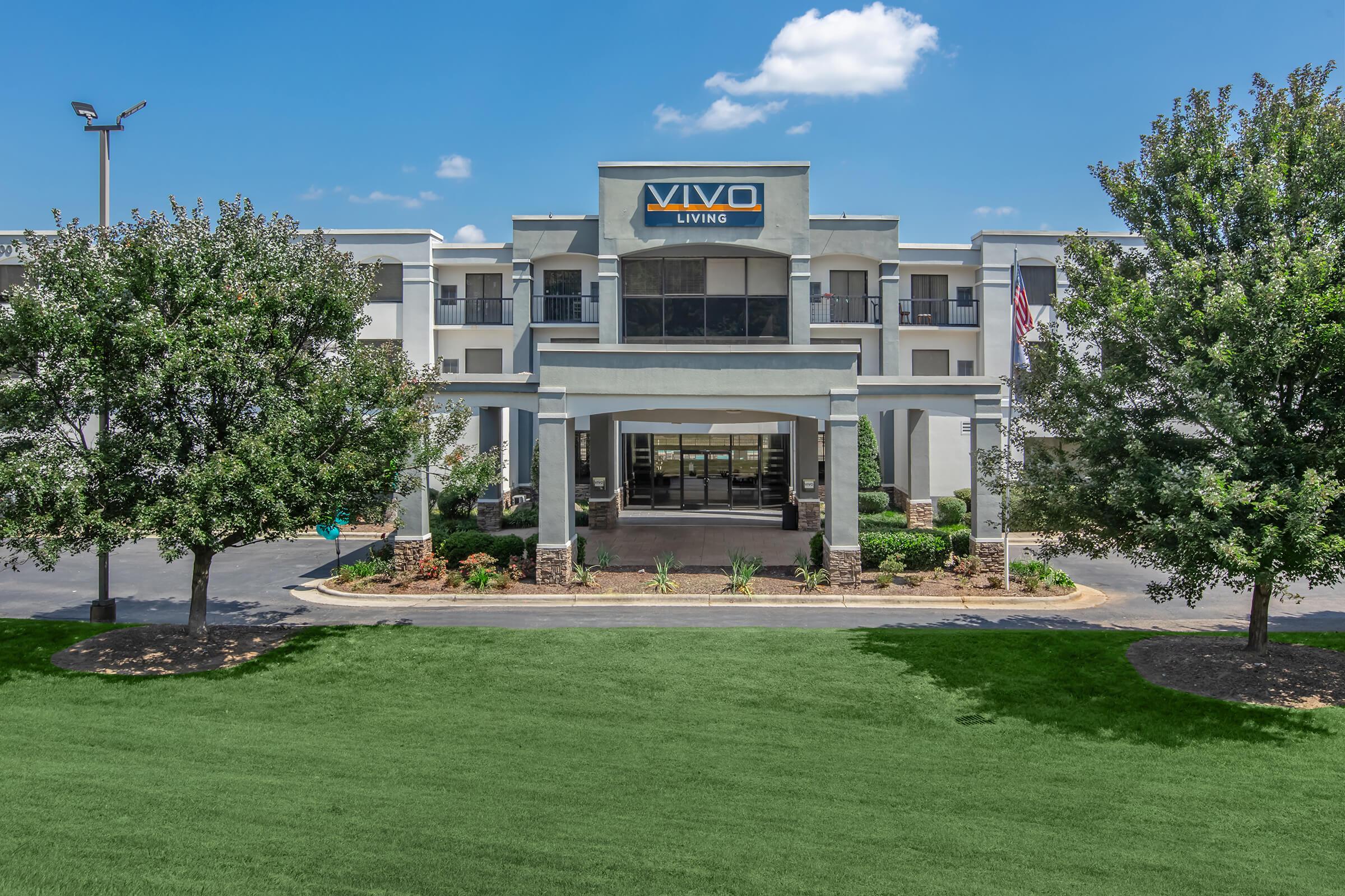 Exterior view of VIVO Living, showcasing a modern building with a prominent entrance, flanked by trees and landscaped areas. The clear blue sky adds to the inviting atmosphere, creating a welcoming impression for potential residents and visitors.