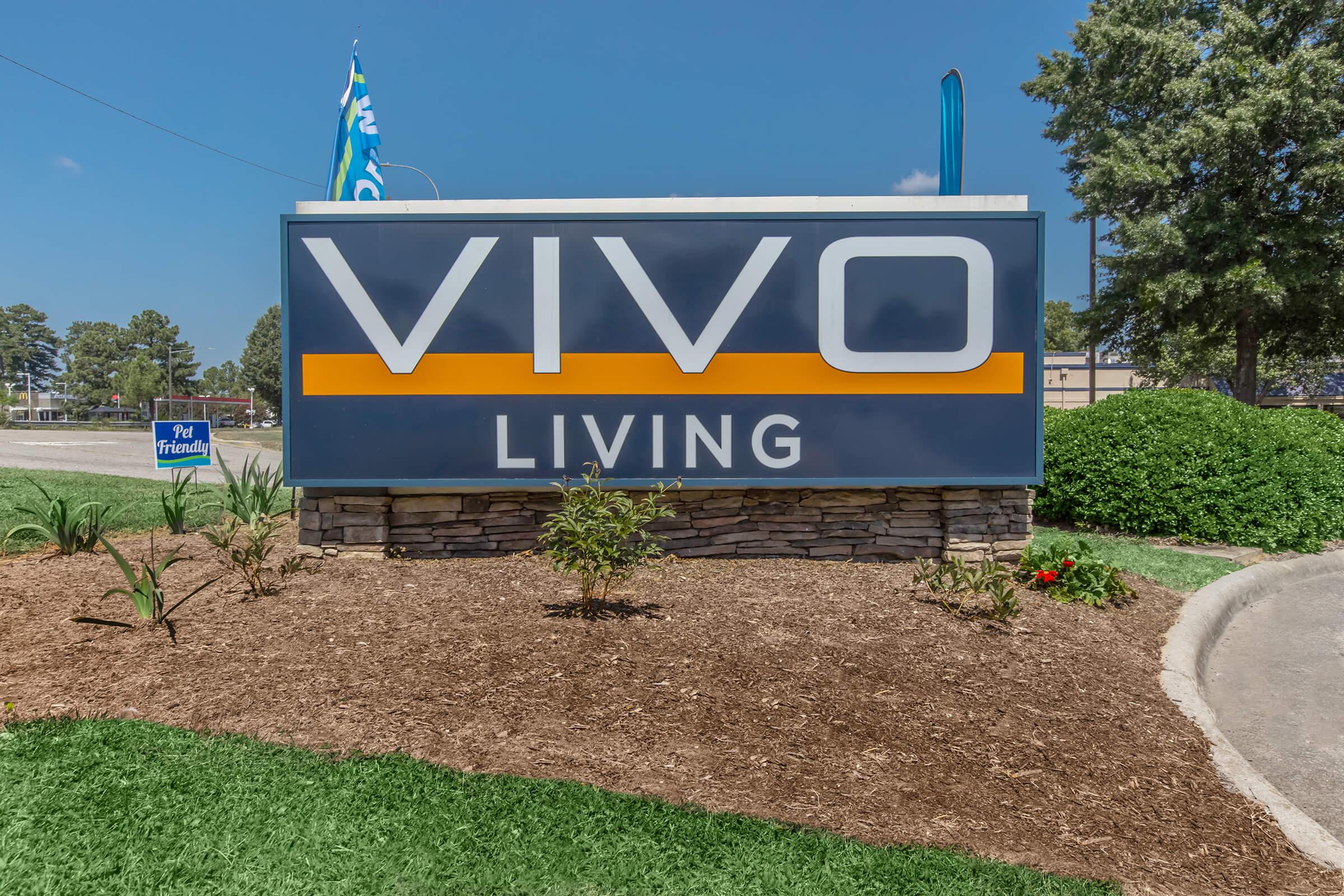 Sign for "VIVO LIVING", featuring bold letters with an orange stripe separating "VIVO" and "LIVING". The sign is surrounded by landscaped greenery, including plants and flowers, against a clear blue sky.