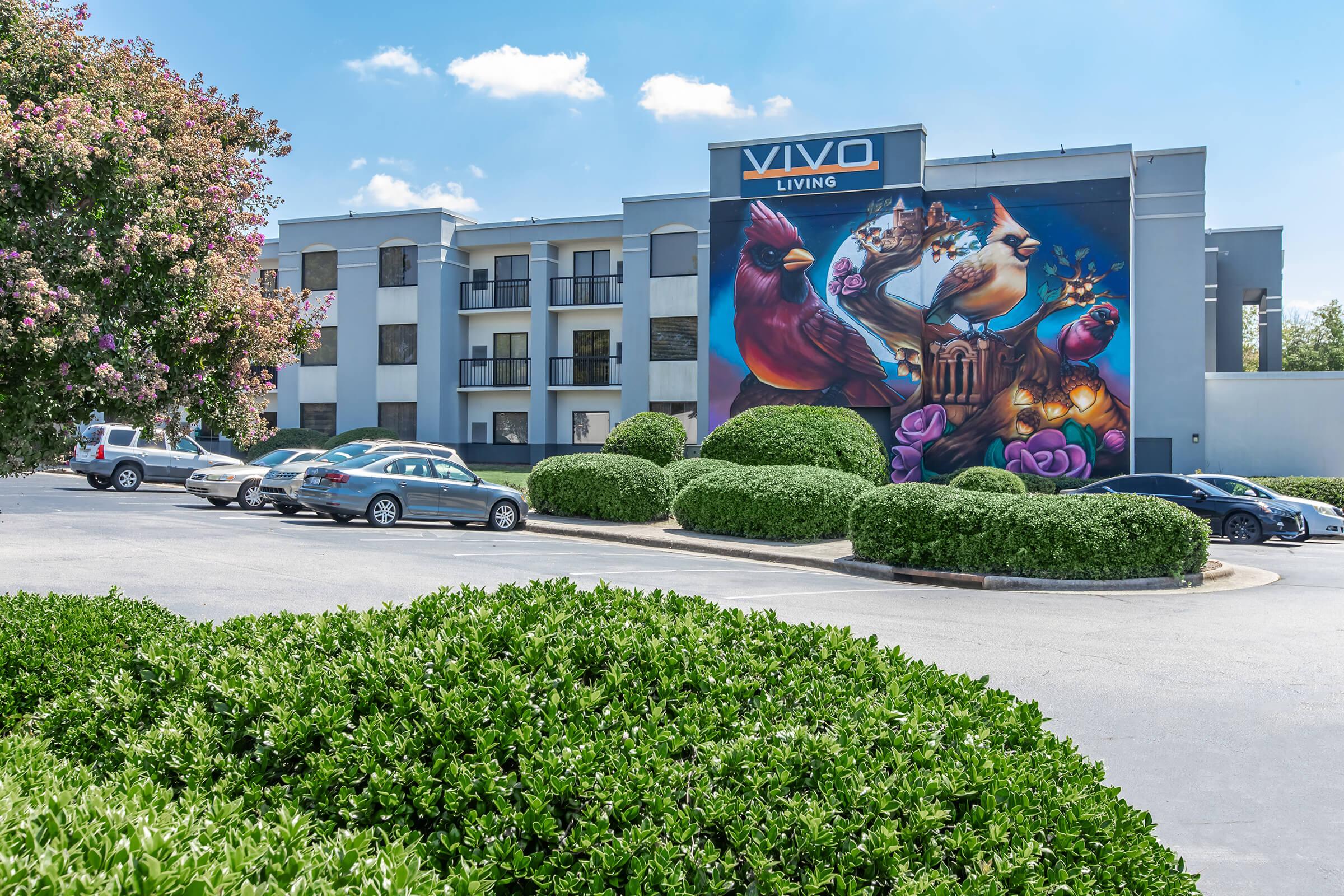 A colorful mural depicting whimsical animals adorns the side of a modern apartment building labeled "VIVO LIVING." The building features several balconies, surrounded by neatly trimmed bushes and a parking area with cars. The sky is bright and blue, creating a lively atmosphere.