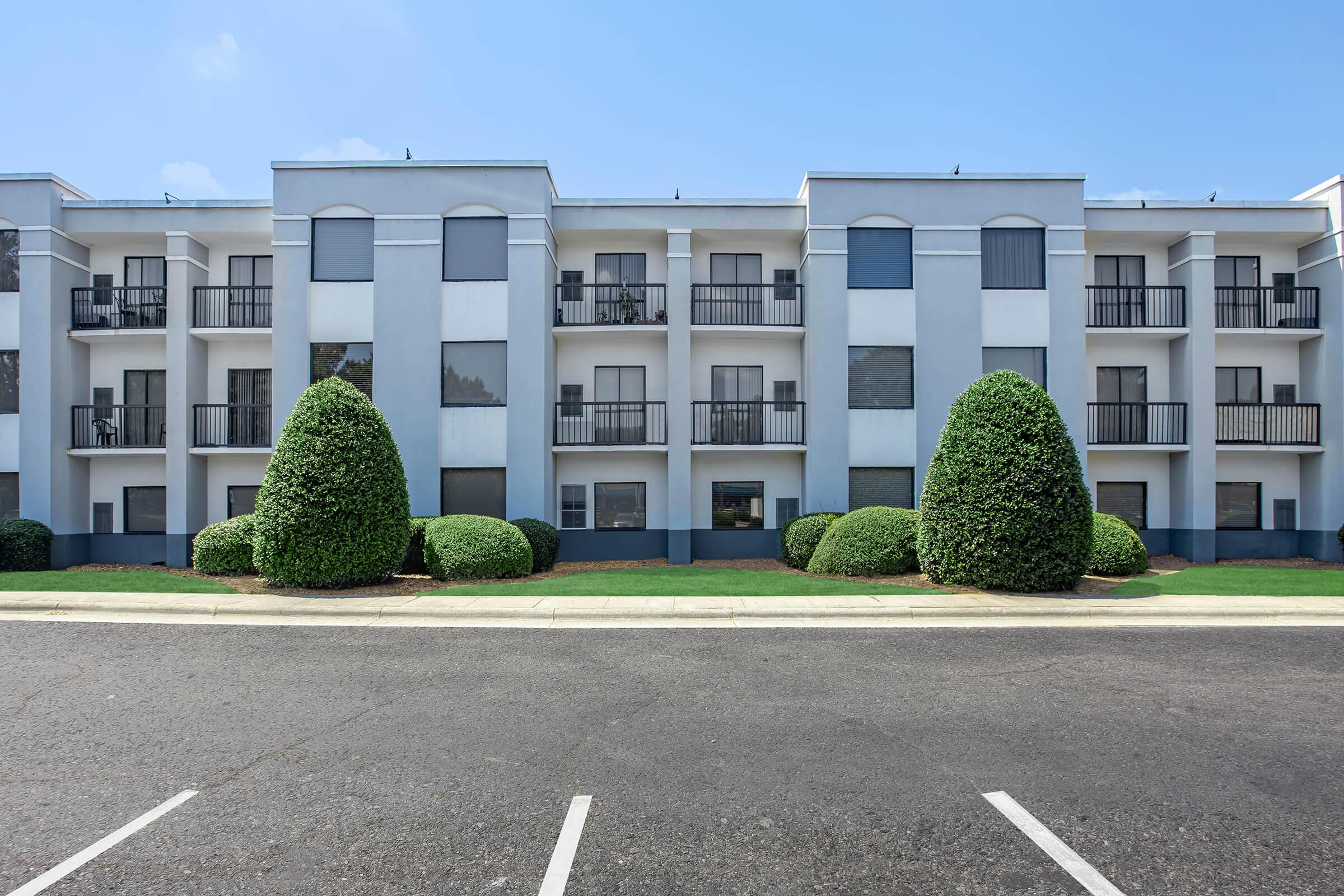 A three-story building with a light-colored facade and dark blue accents. The structure features several balconies with black railings. In front of the building, there are two neatly shaped green bushes and a paved parking area with empty parking spaces. The sky is clear and blue.