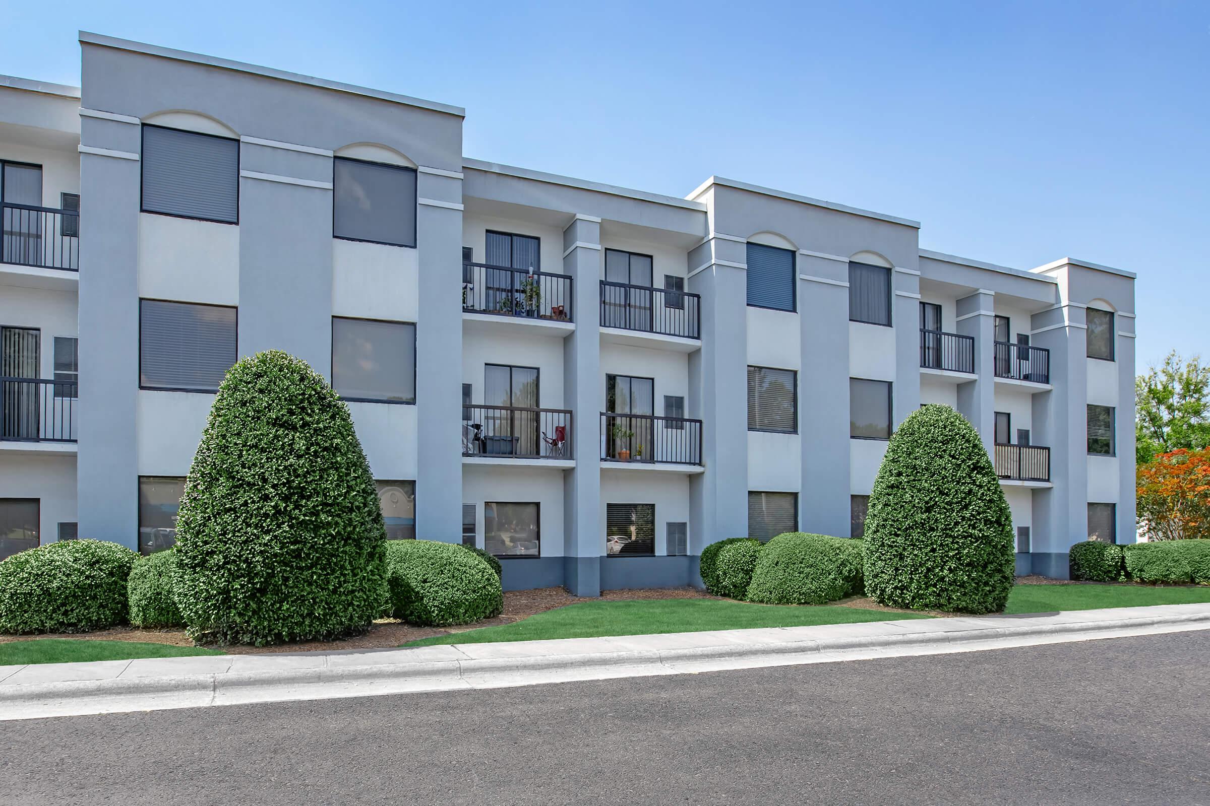 Modern multi-story apartment building featuring a light gray façade, large windows with dark screens, and decorative hedges. The building has balconies on each floor and is situated along a paved road, with green grass and colorful trees in the background under a clear blue sky.