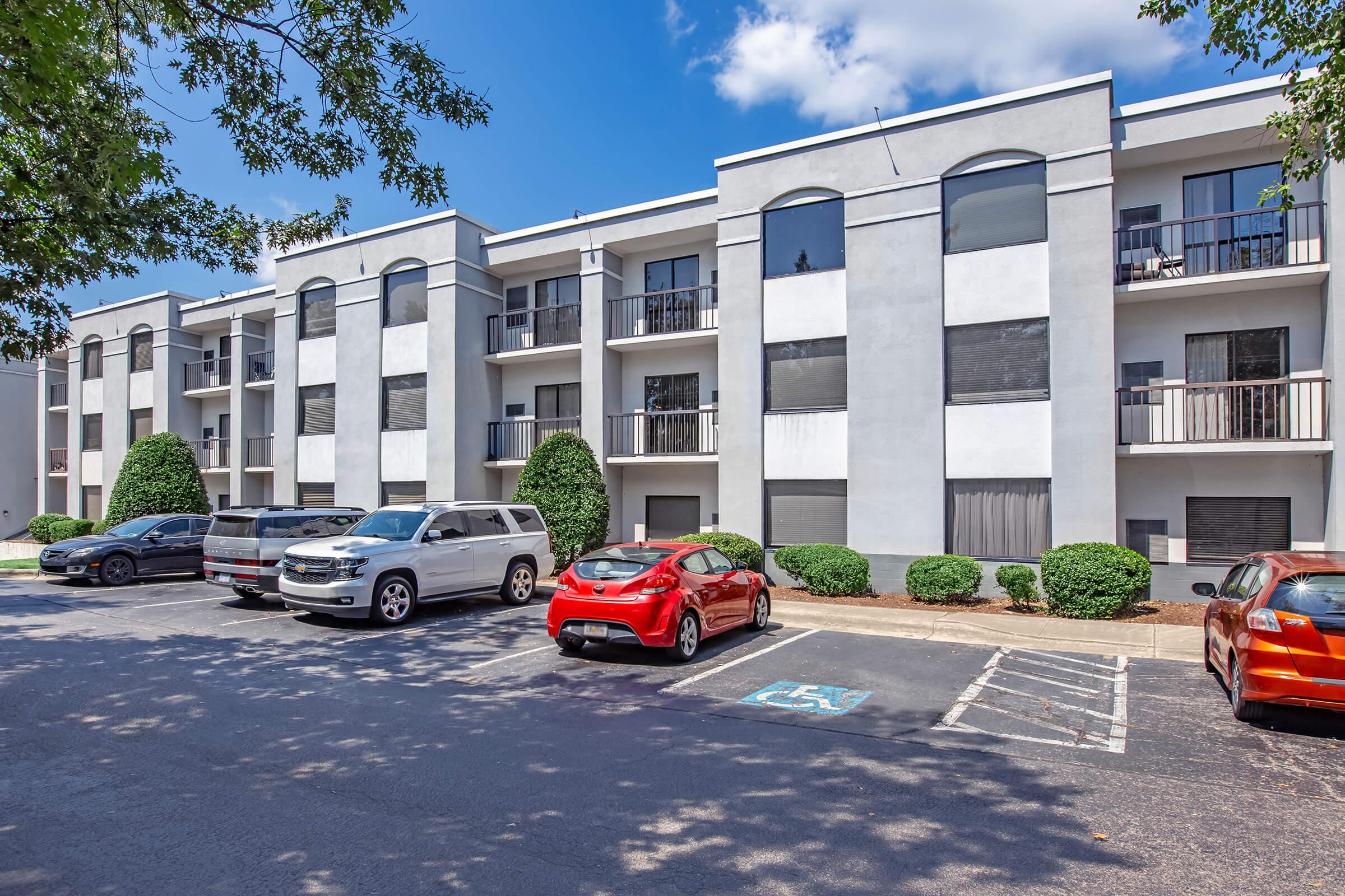 A modern multi-unit apartment building with three stories, featuring balconies and large windows. The parking area in front includes several vehicles, including a red car and an SUV. Surrounding the building are well-maintained shrubs and trees, under a clear blue sky.