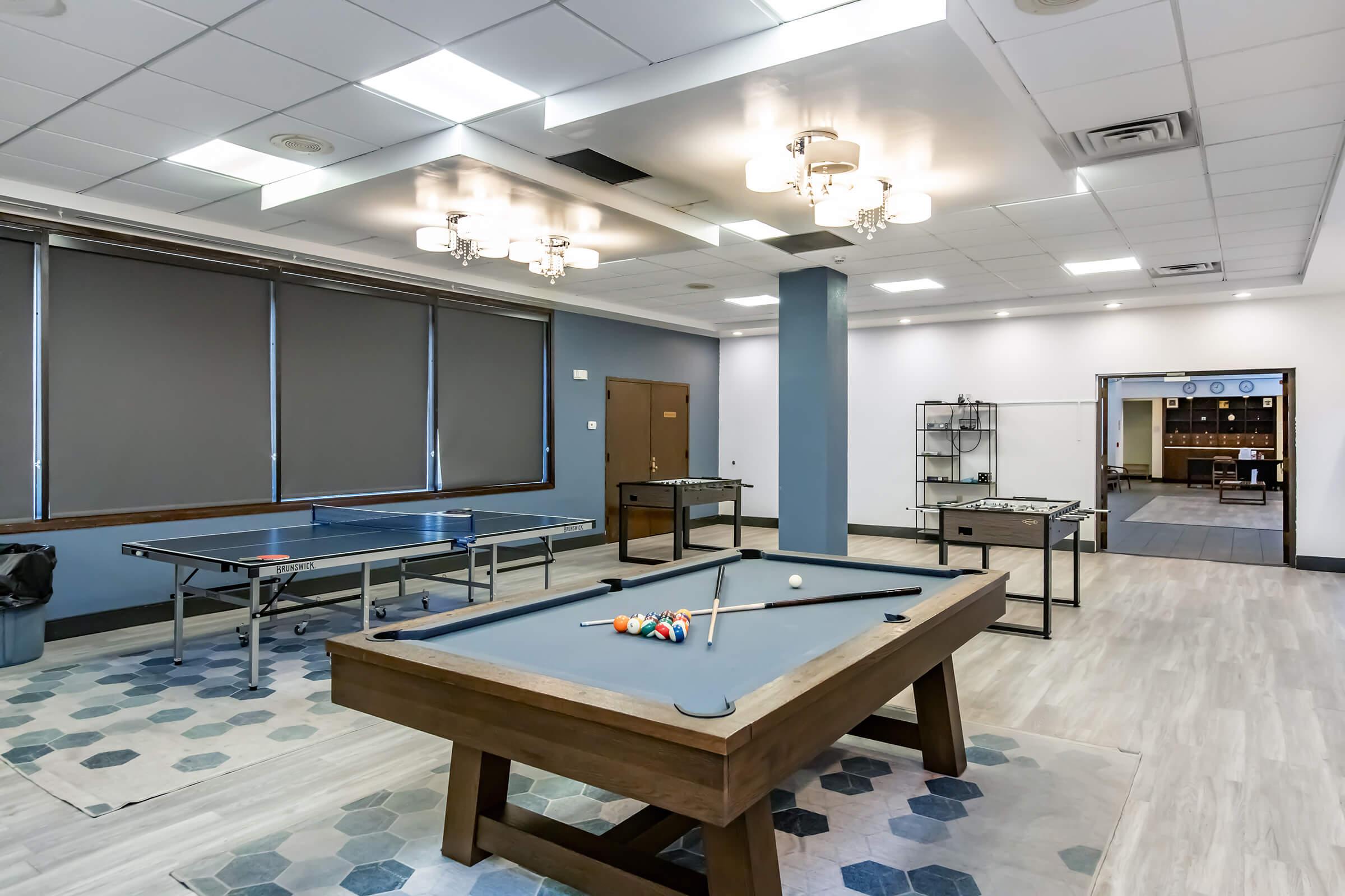 A recreational room featuring a pool table in the foreground, a ping pong table on the left, and several game tables in the background. The room has modern lighting fixtures, grey walls, and a stylish floor design, creating a welcoming atmosphere for leisure activities.
