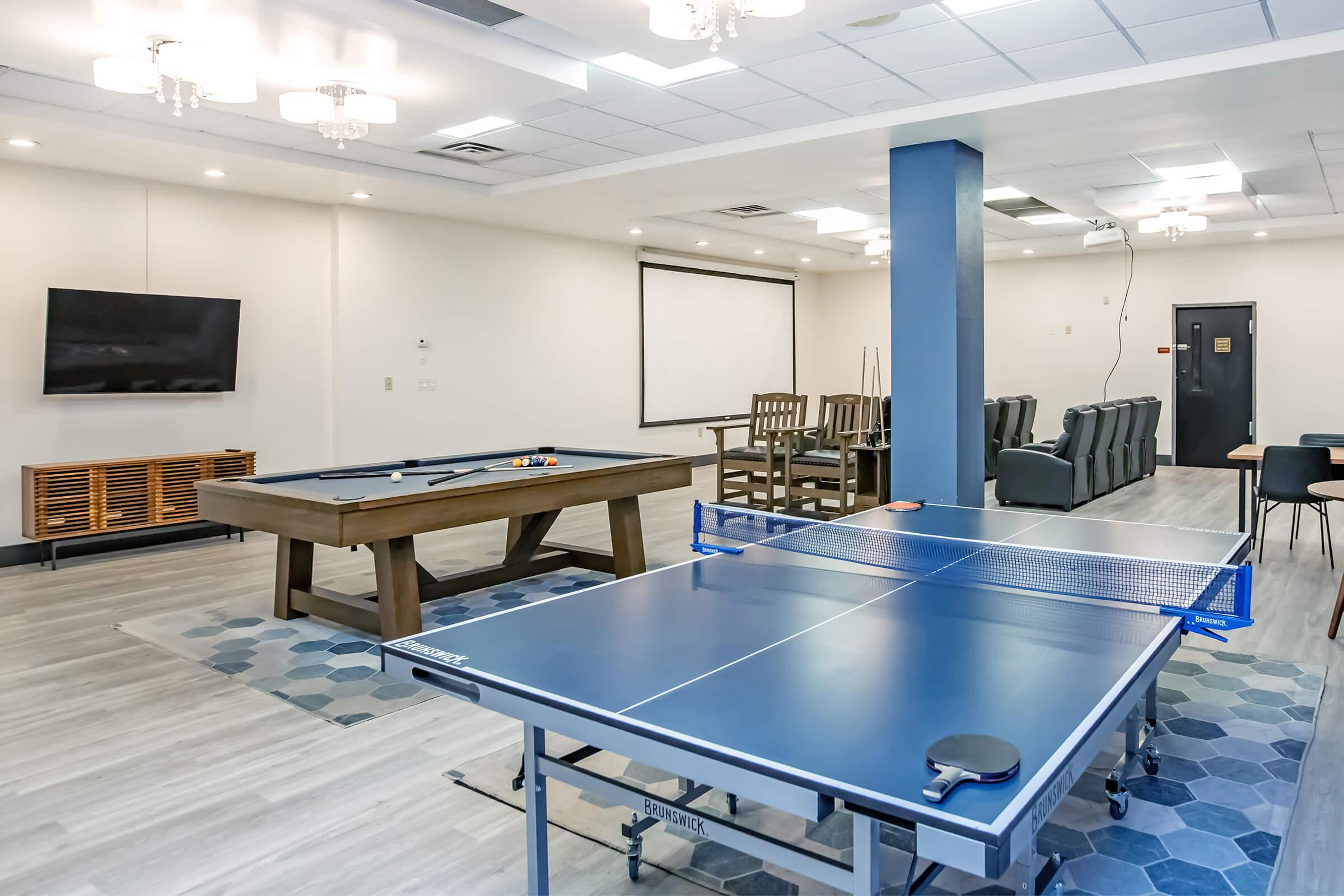 A brightly lit recreational room featuring a ping pong table, a pool table, and seating areas. The walls are painted light colors, with a large television mounted on one side. Modern chandeliers hang from the ceiling, creating a welcoming atmosphere for social and leisure activities.