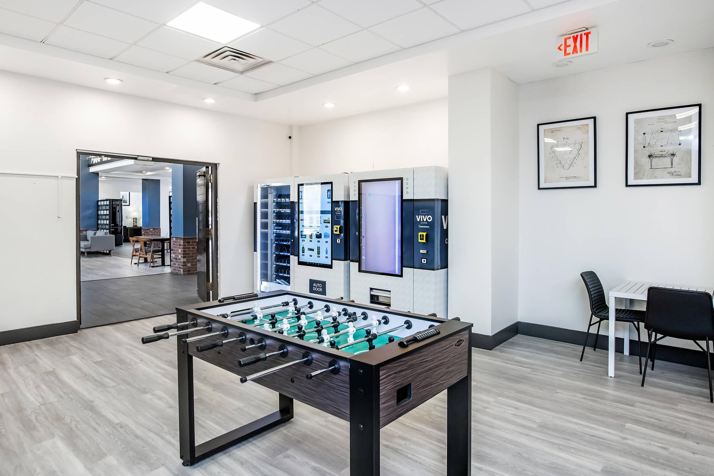 A foosball table in a modern, brightly lit room. In the background, vending machines and a table with chairs are visible. The décor features a clean, minimalist design with neutral colors, and framed artwork on the walls. Subtle natural light enhances the inviting atmosphere.