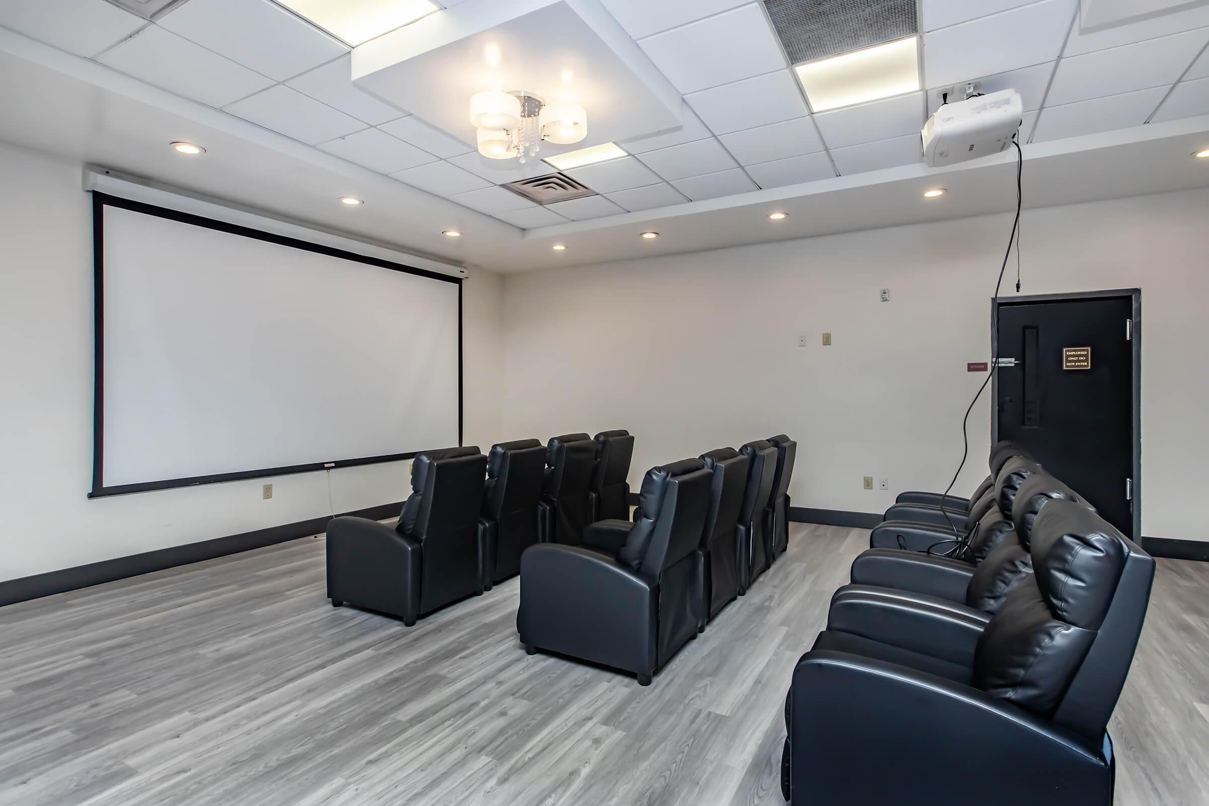 A small room with a large projection screen at the front. There are multiple dark leather recliner chairs arranged in rows facing the screen. The floor has a wooden appearance, and ceiling lights provide illumination. A projector hangs from the ceiling, and the walls are painted in neutral colors.