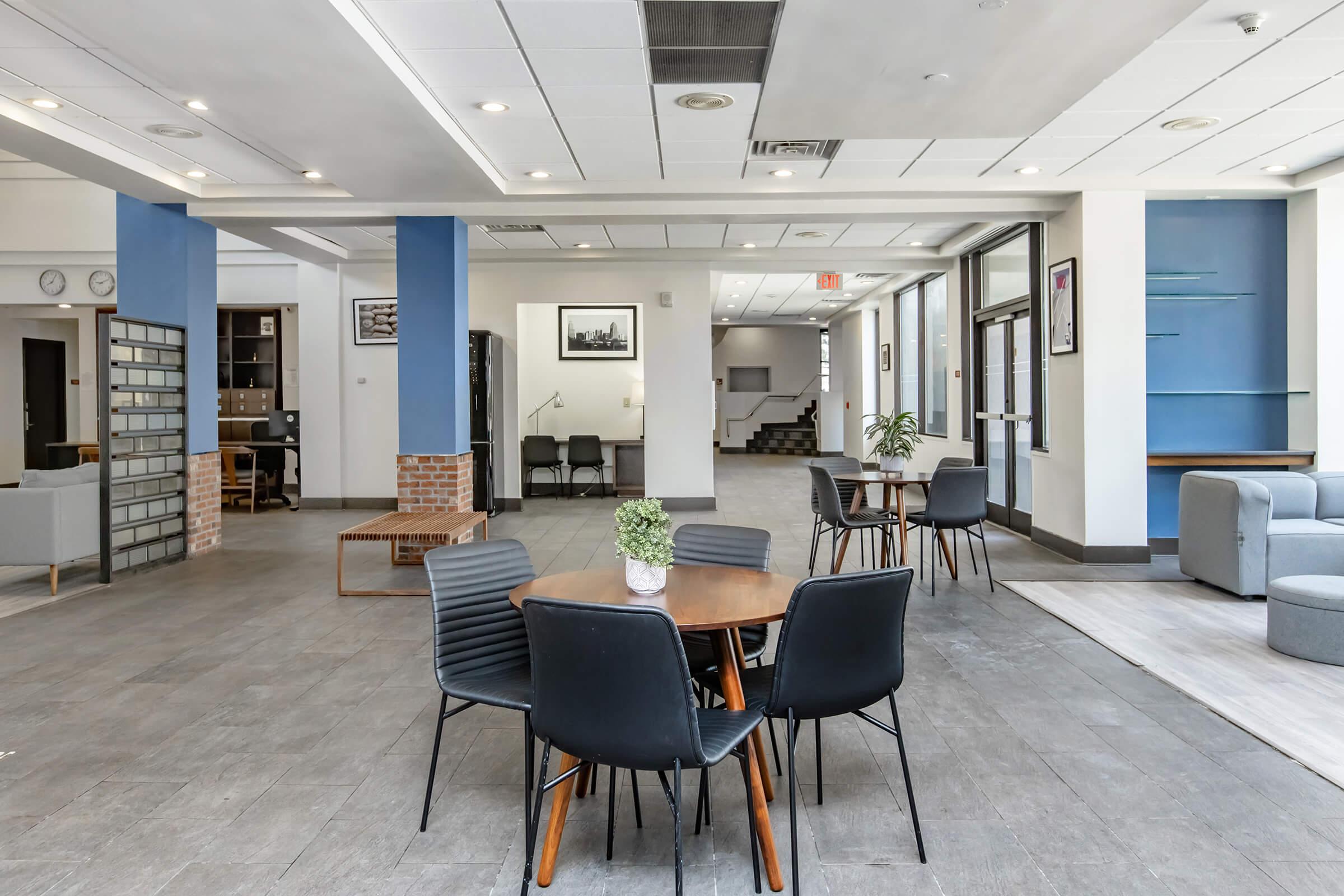 A spacious and modern interior showcasing a common area with several black chairs and wooden tables. The room features large windows, contemporary furnishings, and blue accent walls. There are seating areas with gray sofas, plants, and a staircase in the background, creating a welcoming atmosphere.