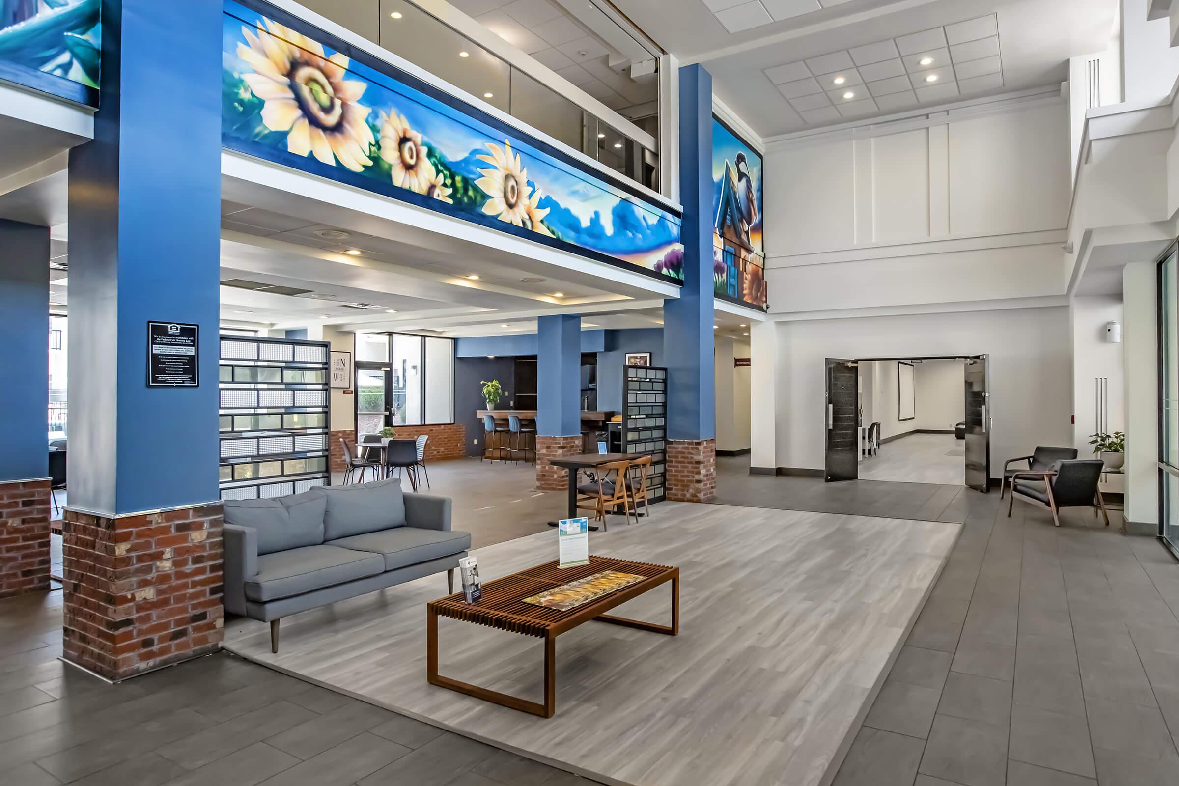A spacious lobby featuring modern decor, large windows, and colorful murals of sunflowers. There is a comfortable gray sofa, a coffee table, and seating areas. The space has a welcoming atmosphere with plants and contemporary furnishings, ideal for relaxation and socializing.