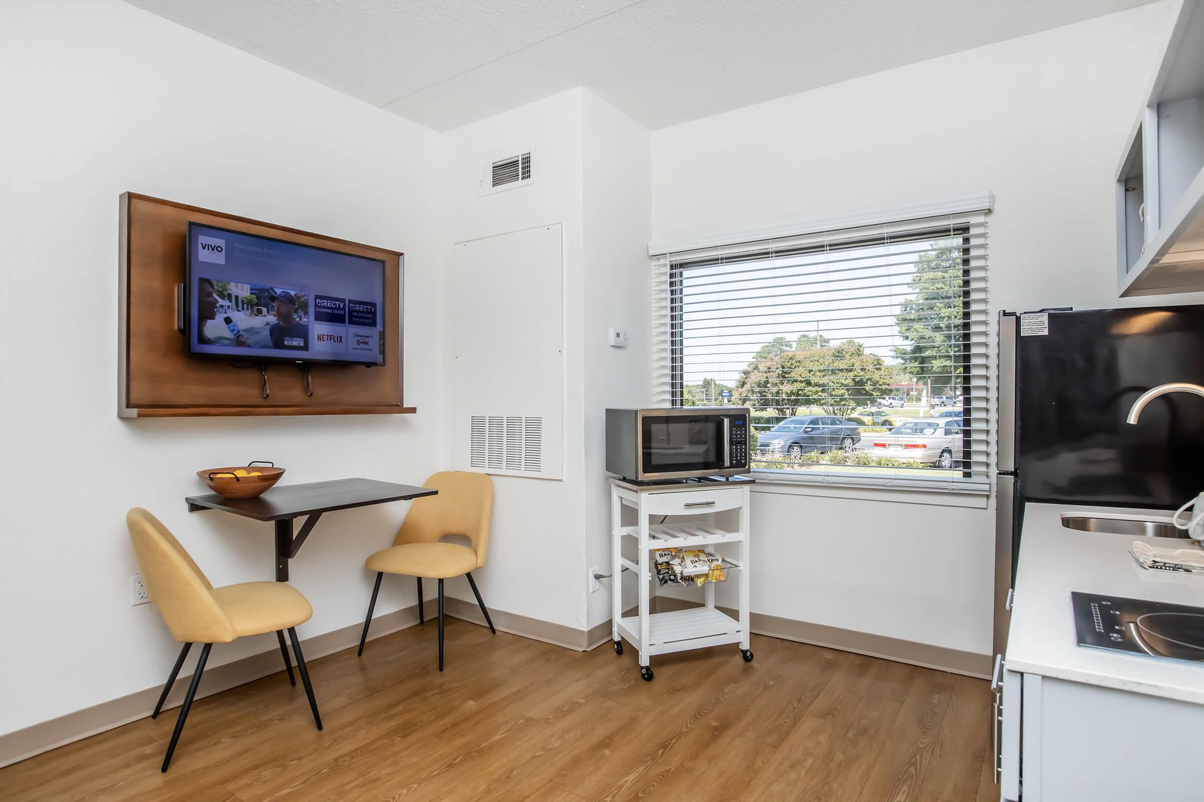 A cozy kitchenette area featuring a wall-mounted TV, a small table with two chairs, a microwave, and a rolling cart. Natural light comes in through a window with blinds, offering a view of the outdoors. The flooring is wood-style, creating a welcoming atmosphere.