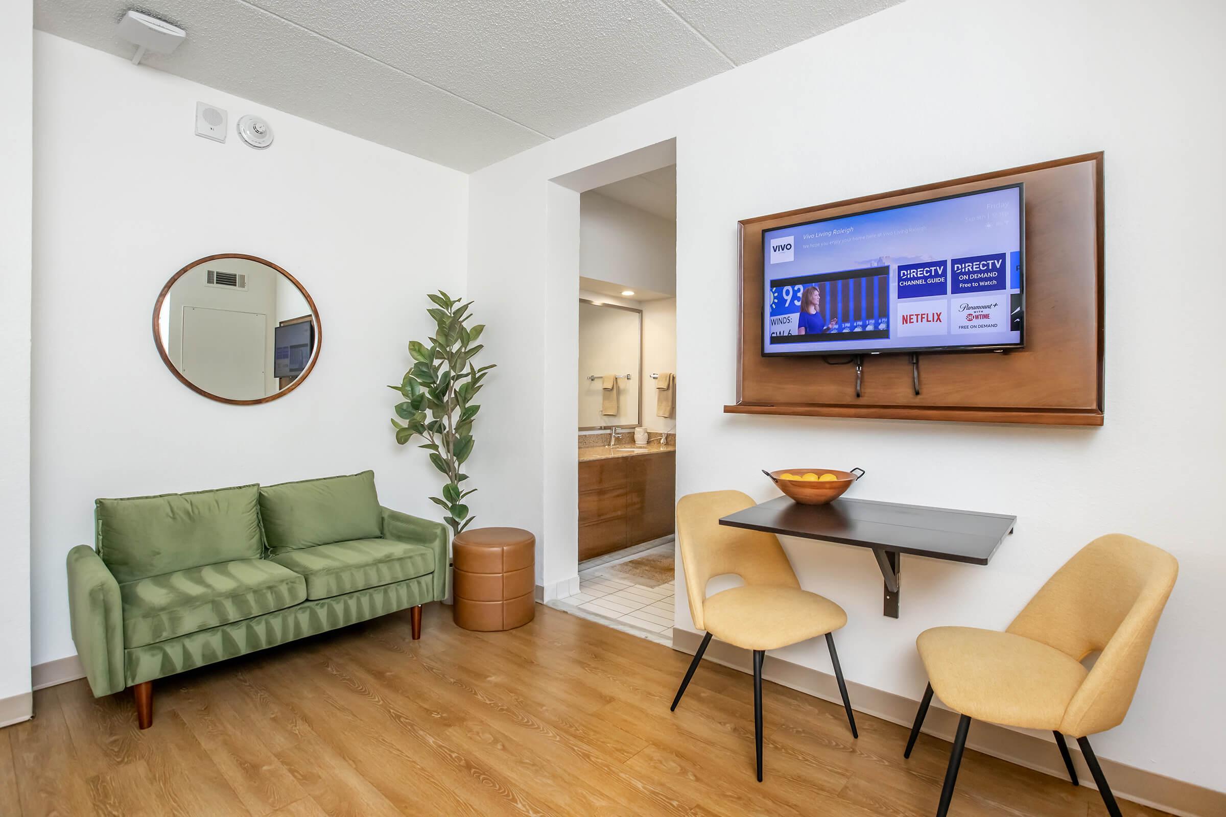 A cozy living space featuring a green sofa, a round mirror on the wall, a small dining table with yellow chairs, and a wall-mounted TV displaying streaming options. A potted plant adds a touch of greenery, and a bathroom is visible in the background. The flooring is wooden and well-lit.