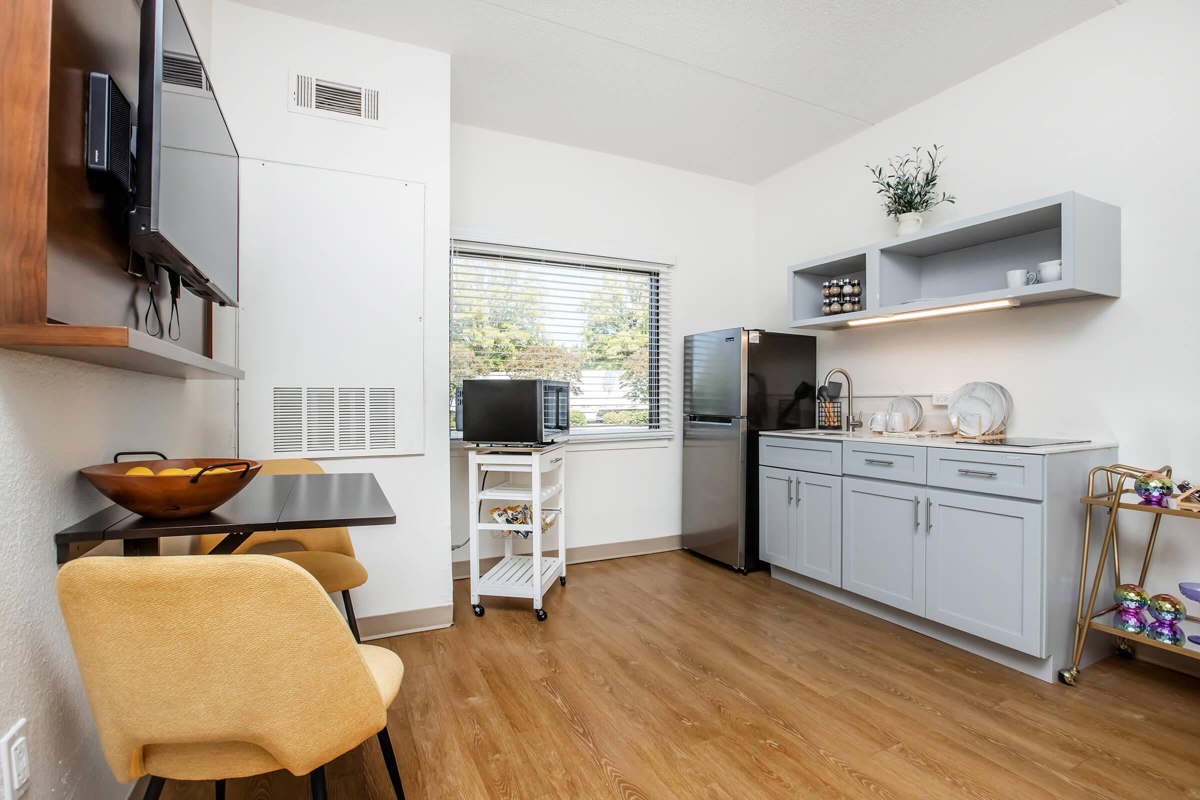 A modern kitchenette featuring gray cabinets, a stainless steel refrigerator, and a microwave. There is a small table with two yellow chairs, a window allowing natural light, and shelves displaying glassware. The flooring is wooden, creating a warm and inviting atmosphere.
