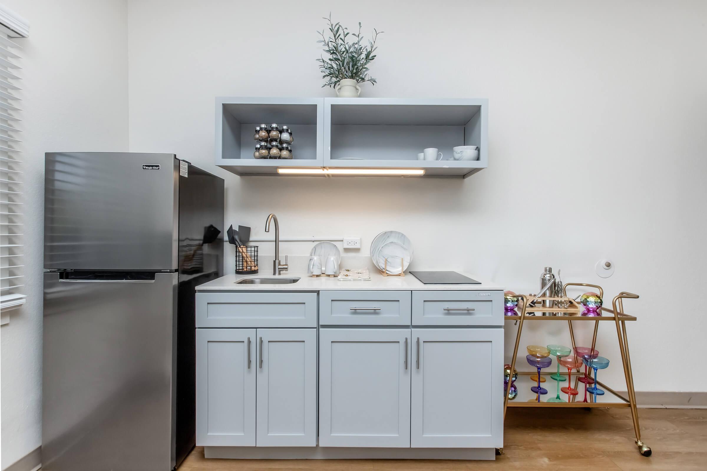 A modern kitchen space featuring a stainless steel refrigerator, light gray cabinets, and an open shelf with decorative items. A countertop has dishware and a small sink, while a stylish bar cart with colorful drinkware is positioned beside it. The walls are plain, contributing to a clean aesthetic.
