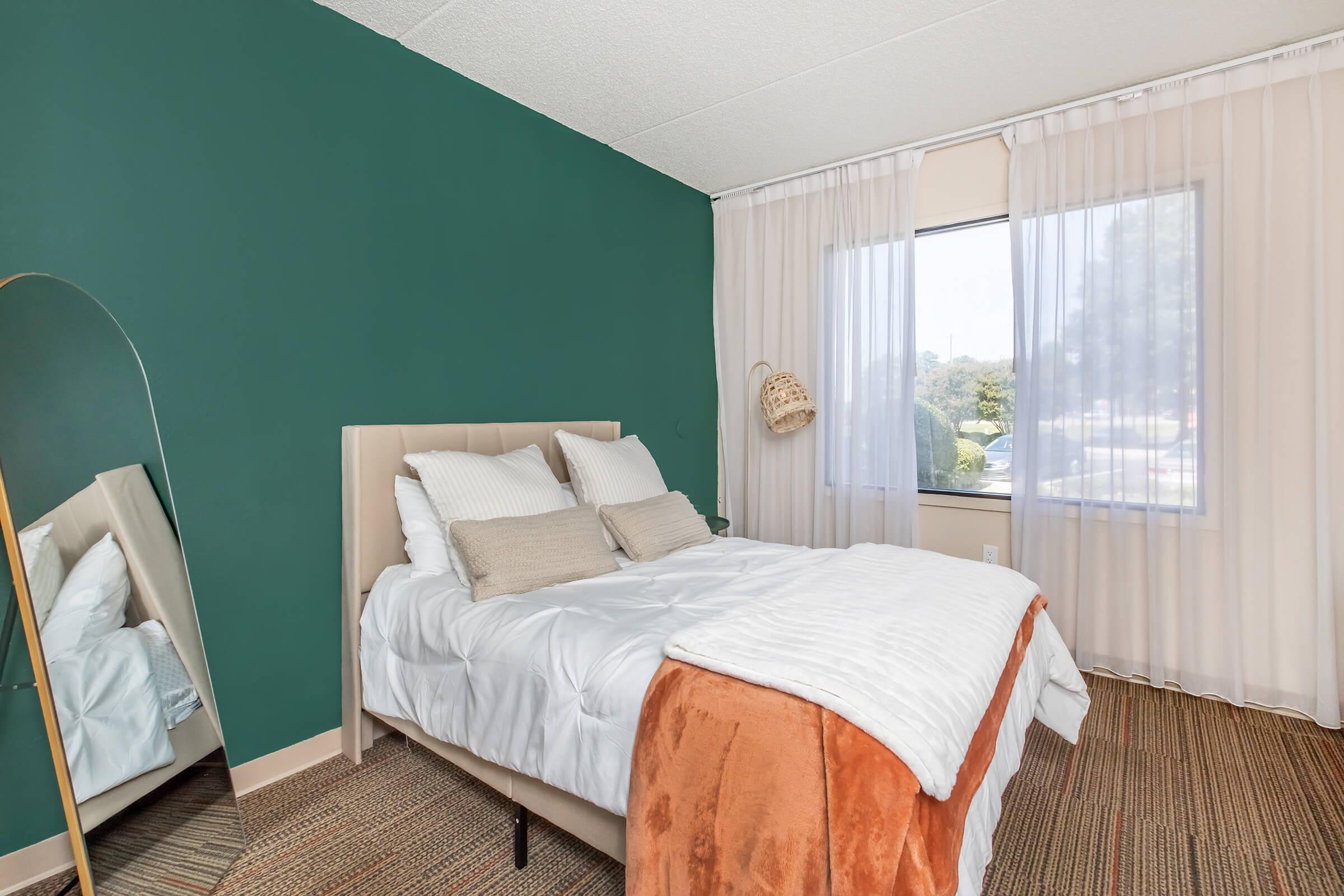 A cozy bedroom featuring a bed with white linens and an orange throw blanket. The walls are painted a rich green, complemented by sheer curtains covering a large window. A full-length mirror stands beside the bed, and a woven basket hangs on the wall, adding a touch of warmth to the space.