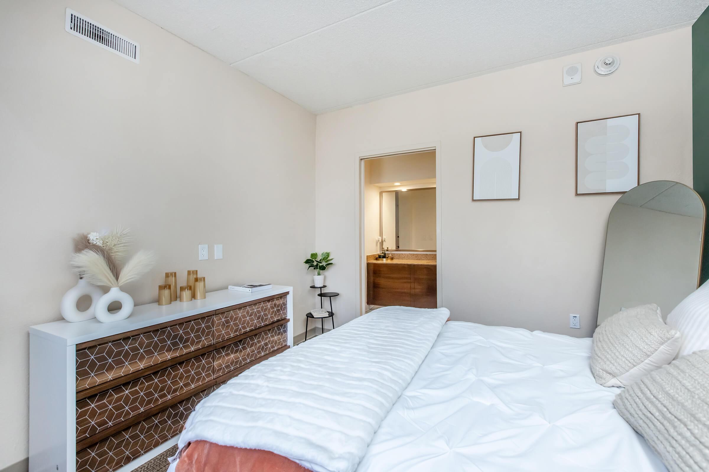 A cozy bedroom with a neatly made bed featuring white linens and a blush-colored throw. There's a decorative dresser with geometric patterns, a small plant, and framed artwork on the walls. A bathroom entrance is visible in the background, adding to the modern and minimalist aesthetic of the space.