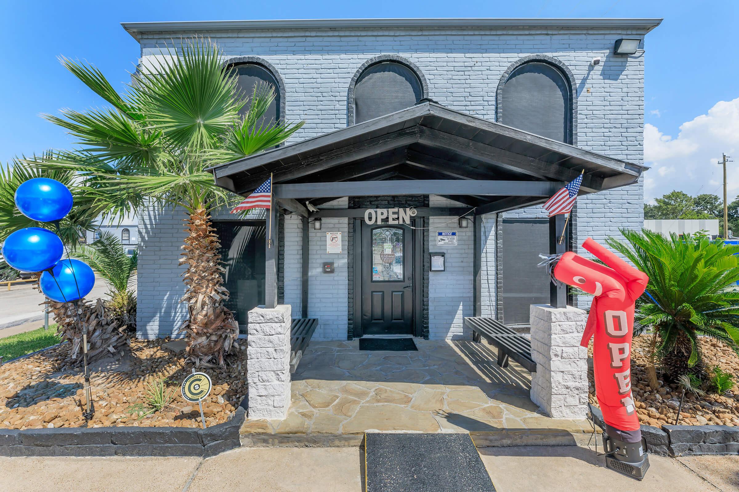 A storefront with a gray brick exterior, featuring a central entrance under a black canopy. On either side of the entrance are palm trees and benches. Blue balloons are tied near the door, and an inflatable “OPEN” sign is waving outside. American flags are displayed, indicating a welcoming atmosphere.