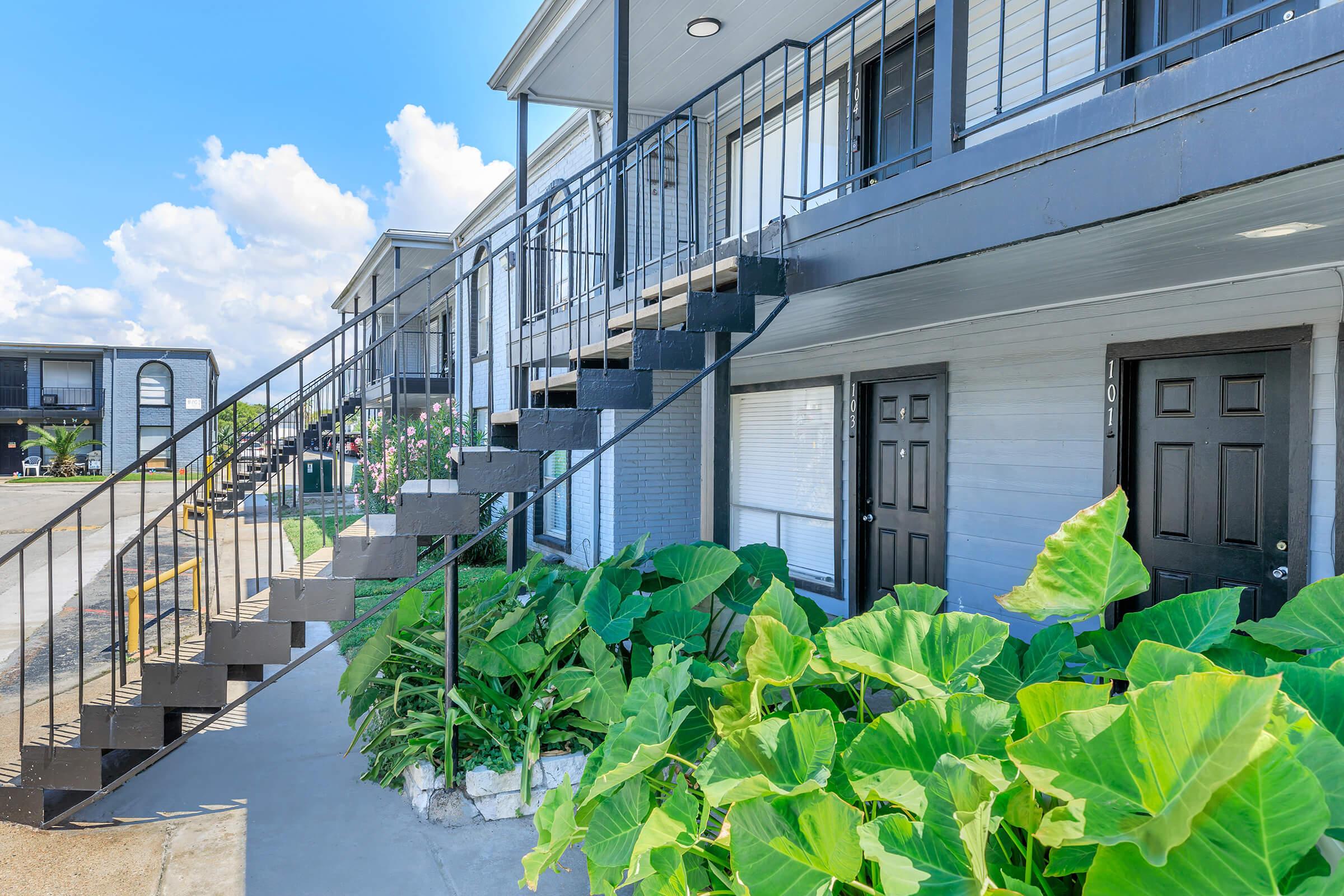 A two-story residential building with a metal staircase leading to the upper floor. The ground level features two dark-colored doors, surrounded by lush green plants. The sky is bright with a few clouds, and the building has a modern aesthetic.