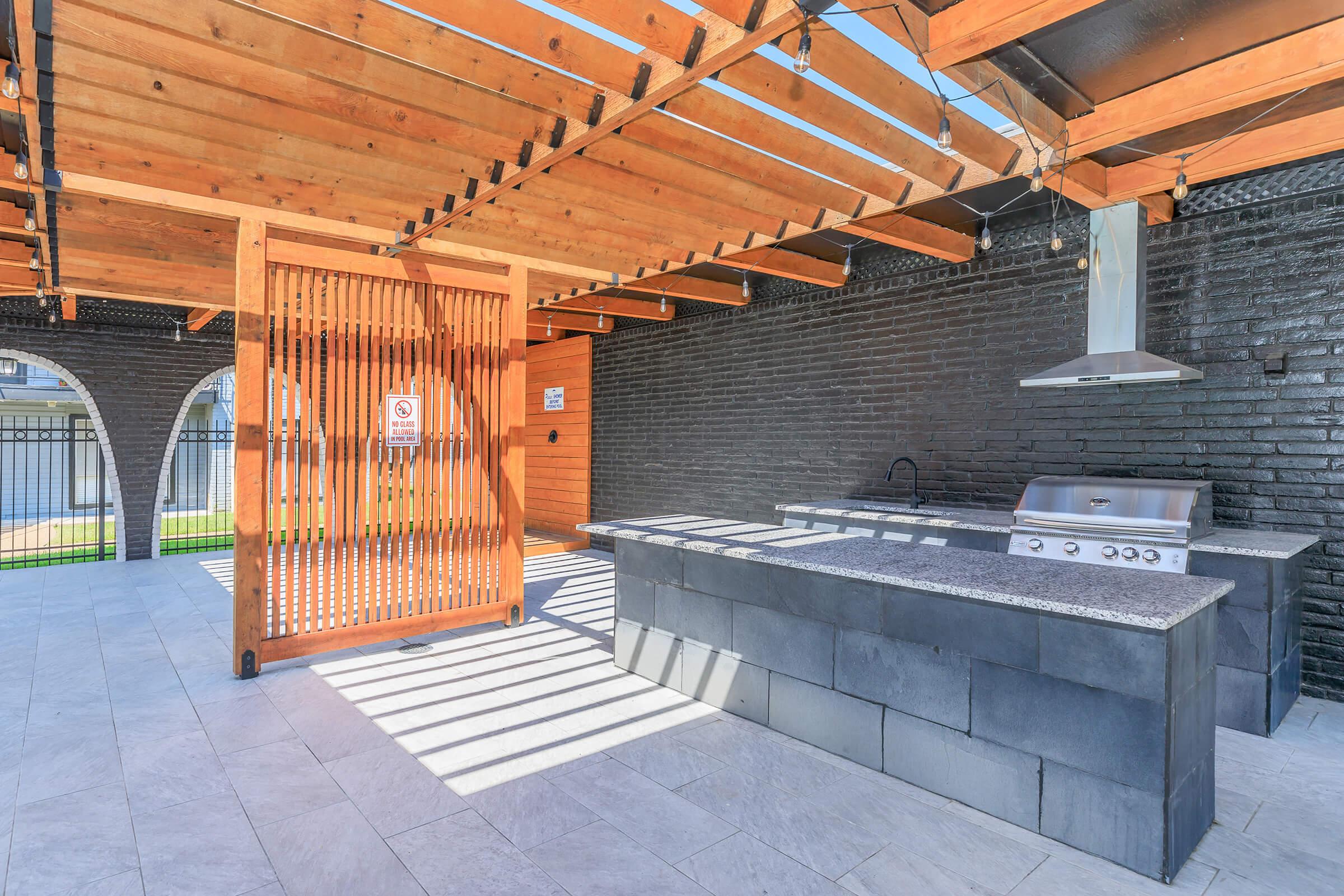Outdoor kitchen area featuring a built-in grill with granite countertop, set beneath a wooden pergola. The space has a modern and open design, with black brick walls and a stone floor, and is illuminated by natural light filtering through the wooden slats above.
