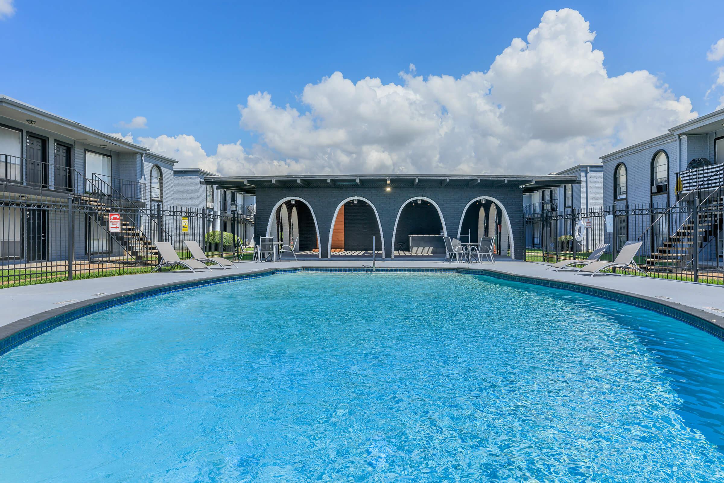 A clear blue swimming pool surrounded by lounge chairs, with an archway and a building in the background. The sky is bright with fluffy white clouds, and there are two two-story buildings on either side of the pool area. Green grass frames the pool, creating a welcoming outdoor space.