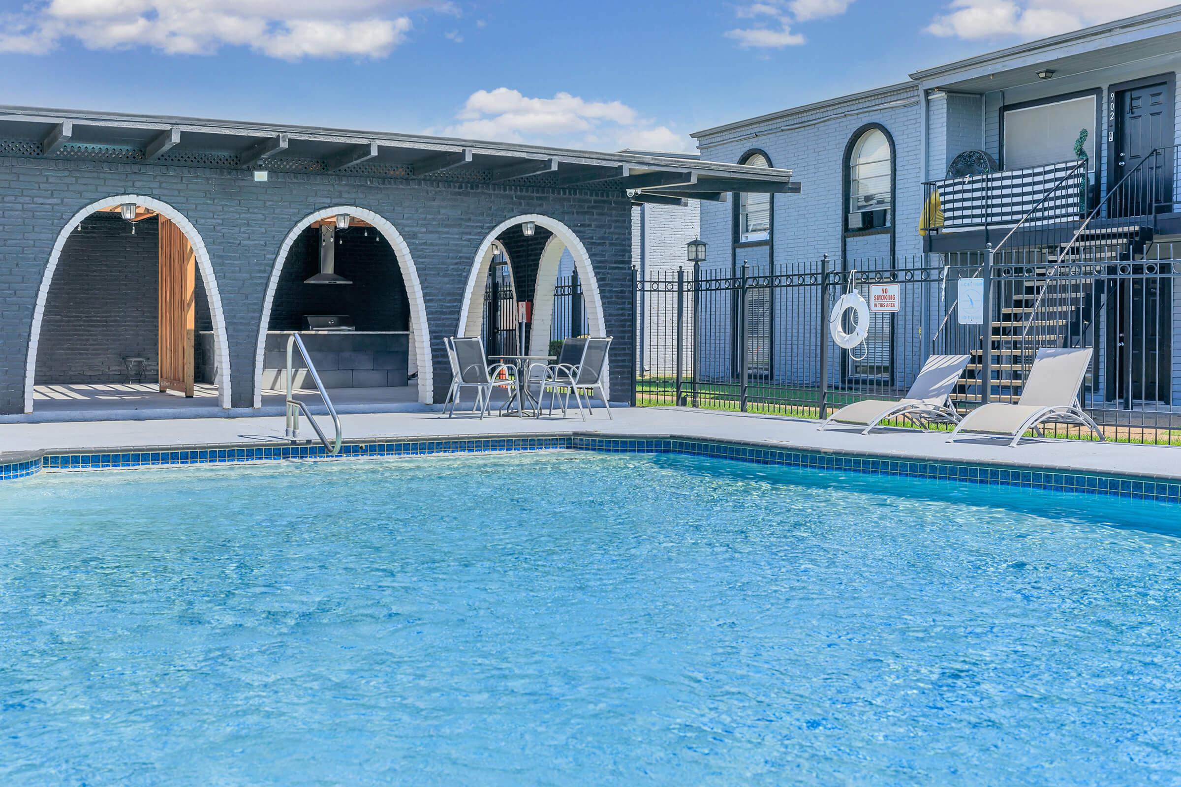 A clear blue swimming pool with a patio area featuring lounge chairs and a covered barbecue space. In the background, a building with multiple levels and a stairway. The scene is sunny with a few clouds in the sky, creating a relaxing outdoor atmosphere.