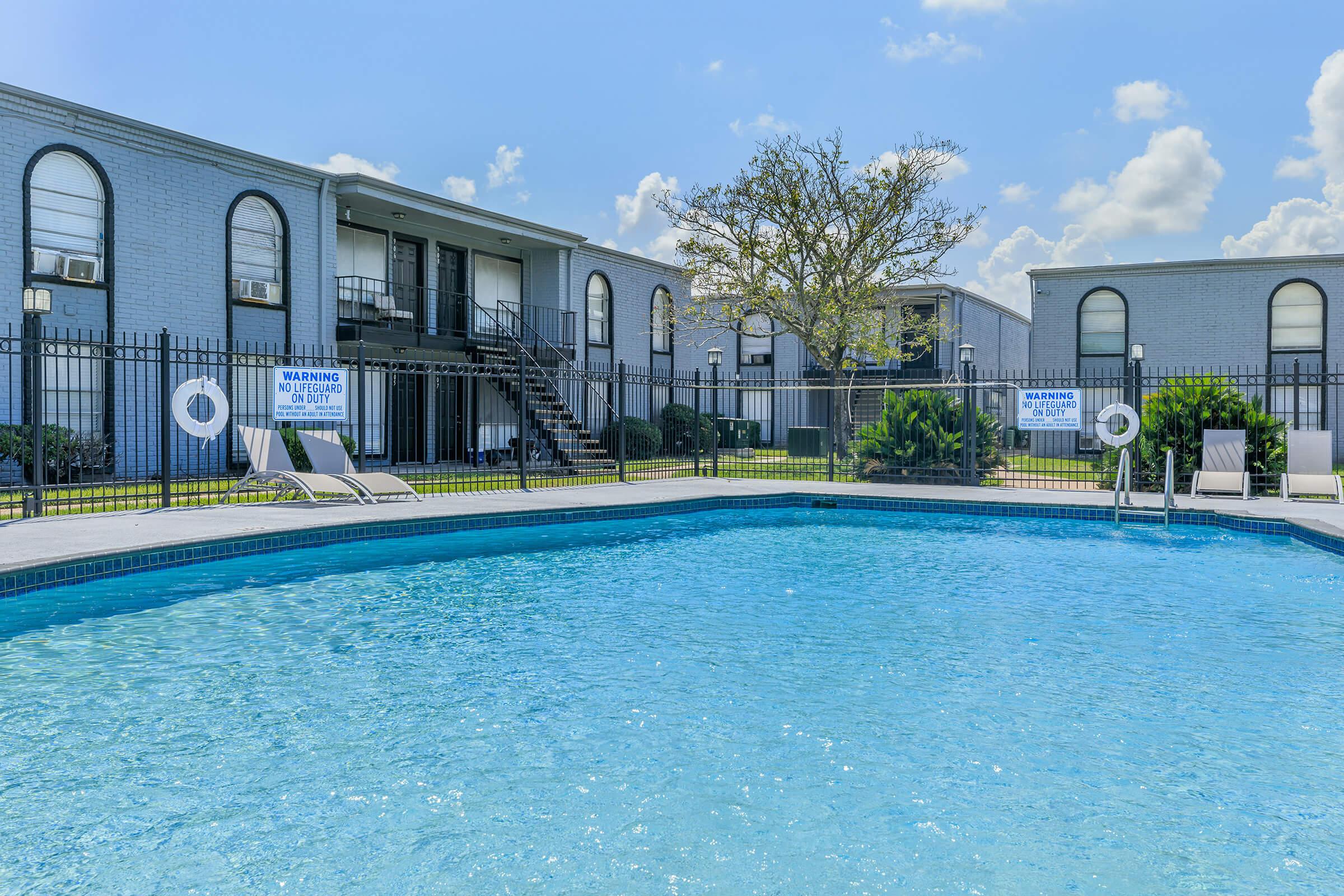 A clear blue swimming pool surrounded by a fence, with lounge chairs on the deck. In the background, two gray residential buildings are visible, along with a tree and bright blue sky with fluffy clouds. Warning signs are posted near the pool area.