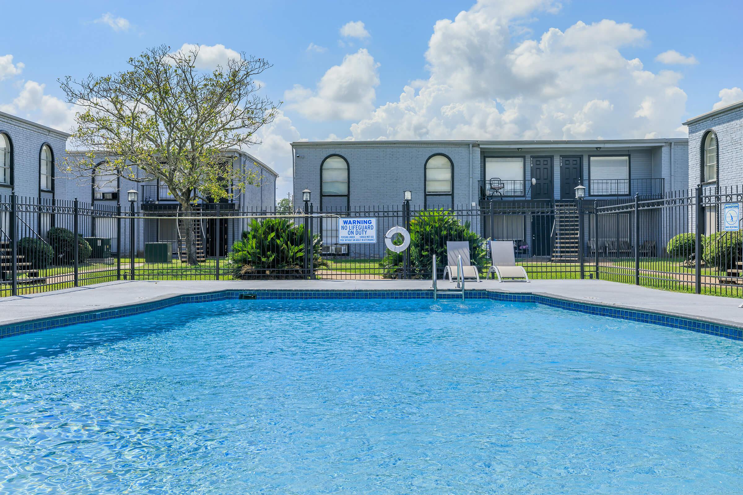 A clear blue swimming pool with a diving board. Surrounding the pool are lush green plants and a fence. In the background, there are two gray apartment buildings with balconies and a sign warning about pool safety. The sky is bright with fluffy white clouds.