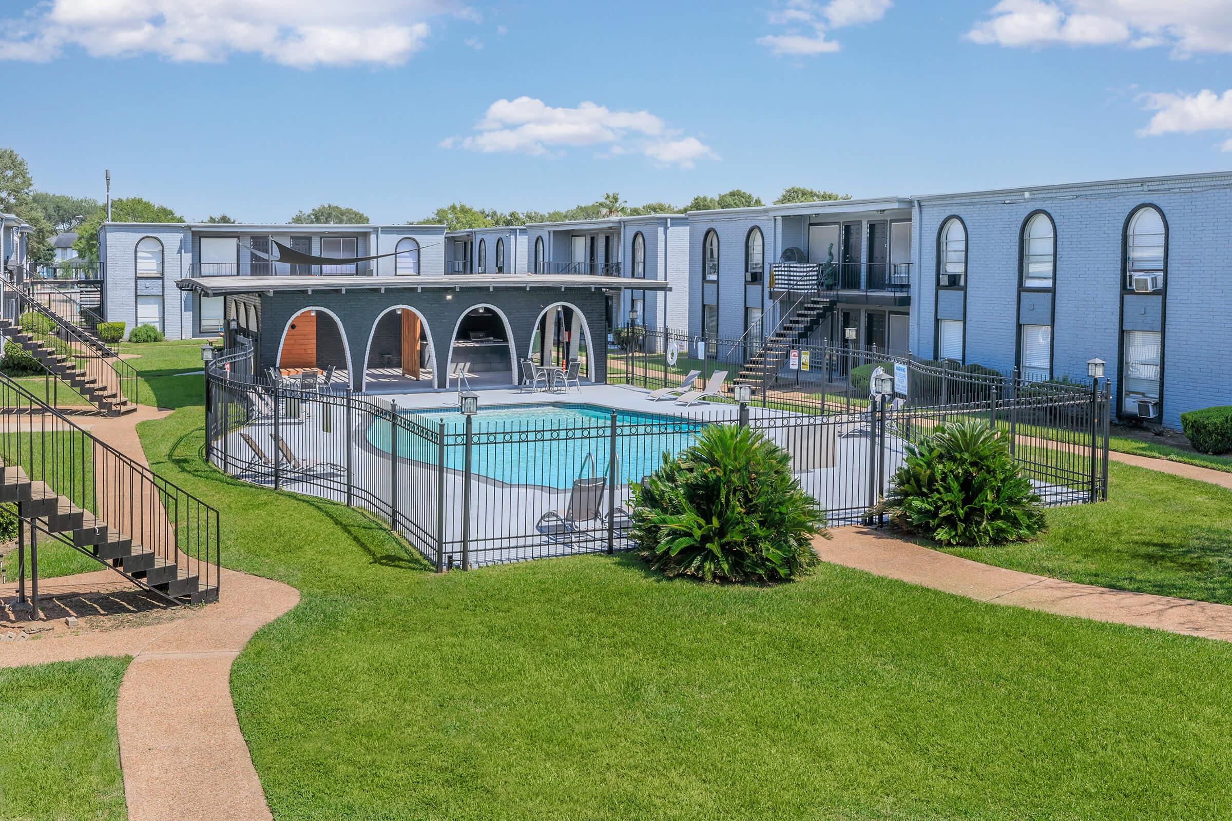 A well-maintained apartment complex featuring a blue swimming pool surrounded by a black fence. Green grassy areas and palm plants are visible, with two sets of stairs leading up to the units. The buildings have arch-shaped windows and balconies, under a clear blue sky with a few clouds.