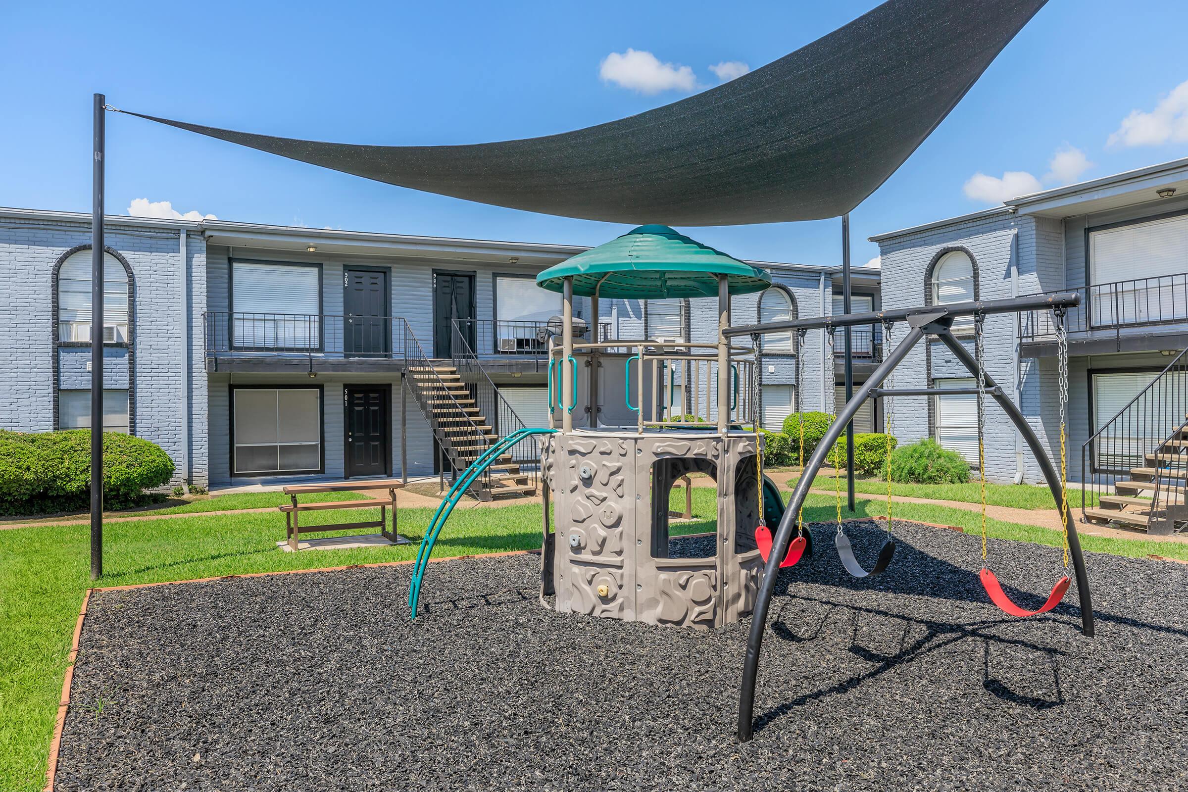 A children's playground featuring a climbing structure with slides and a swing set, situated in an outdoor area surrounded by apartment buildings. The playground is covered by a large shade sail, with green grass and black rubber mulch underneath. Bright blue sky with a few clouds visible.