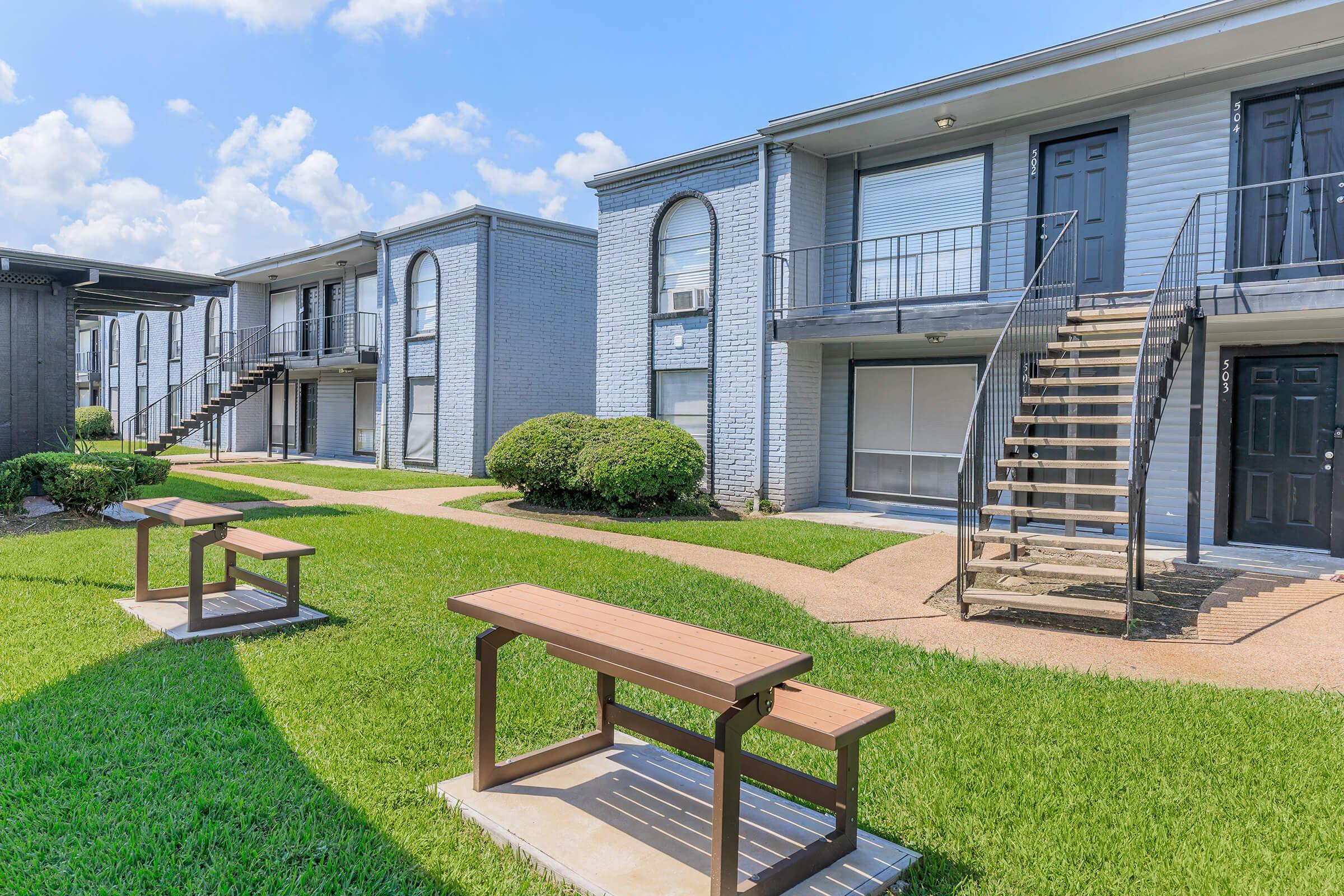 A view of an apartment complex featuring two buildings with stairs leading to second-floor units. Well-maintained grassy areas with benches are placed in front of the buildings, under a clear blue sky with scattered clouds.