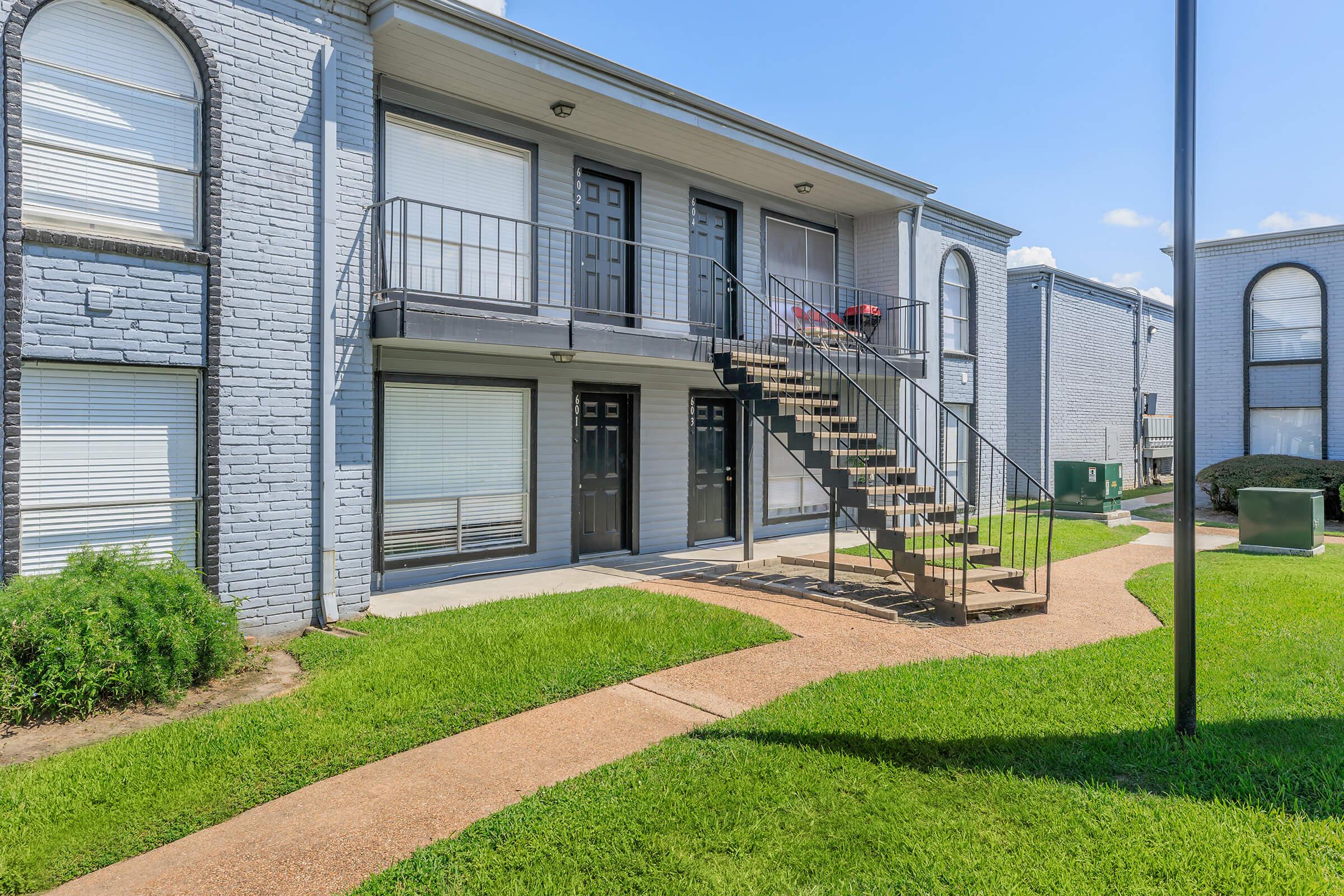 A two-story apartment building with a staircase leading to the upper level. The exterior features grey brick walls and large windows with white blinds. A manicured lawn and pathway are visible in front of the building, along with a green utility box. The sky is clear and blue, indicating a sunny day.