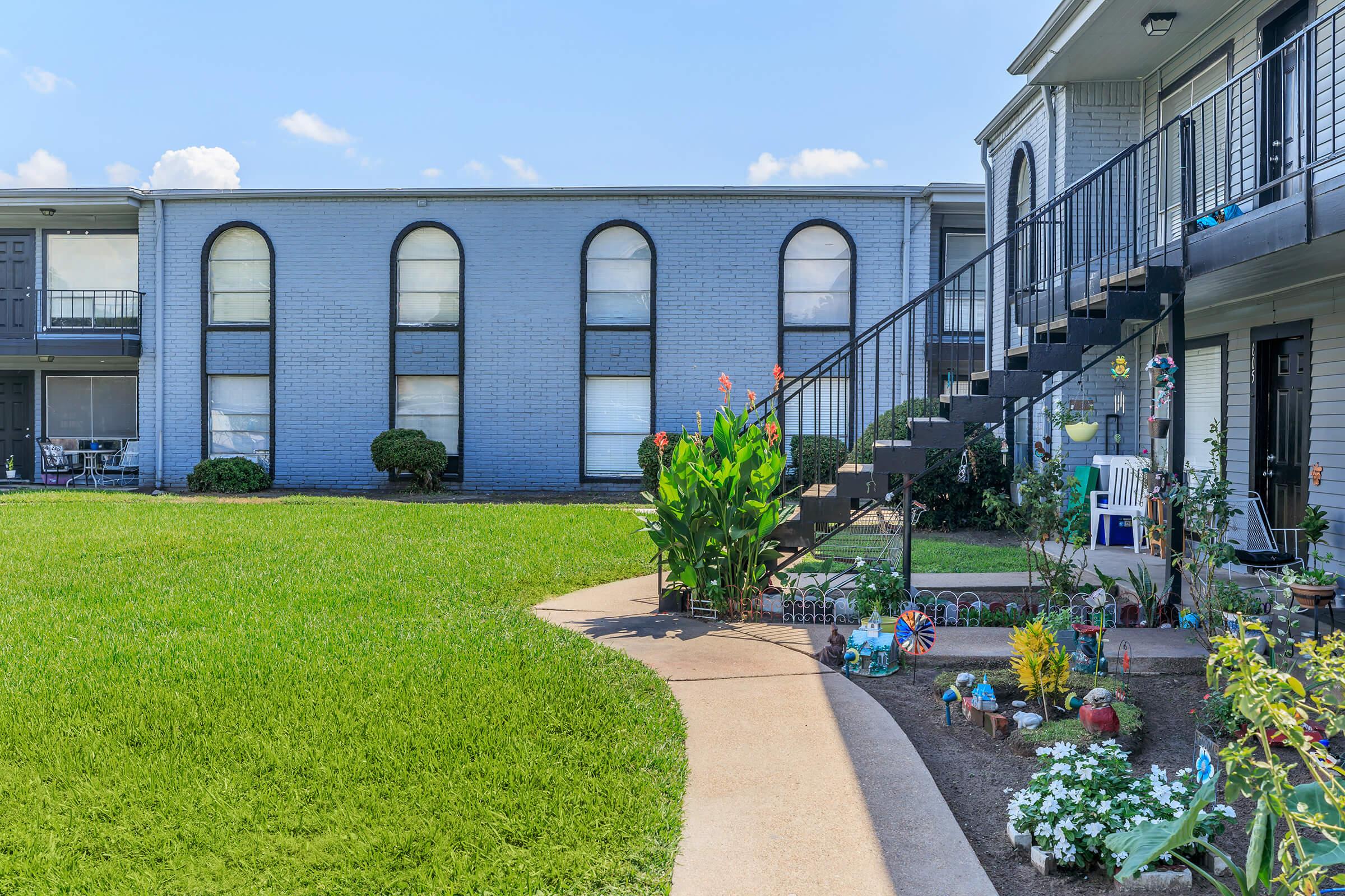 A landscaped courtyard featuring a curved sidewalk leading to an exterior staircase. Vibrant flowers and plants line the path and garden area, with a backdrop of a light blue building and several arched windows. The scene is bright and sunny with a clear sky.