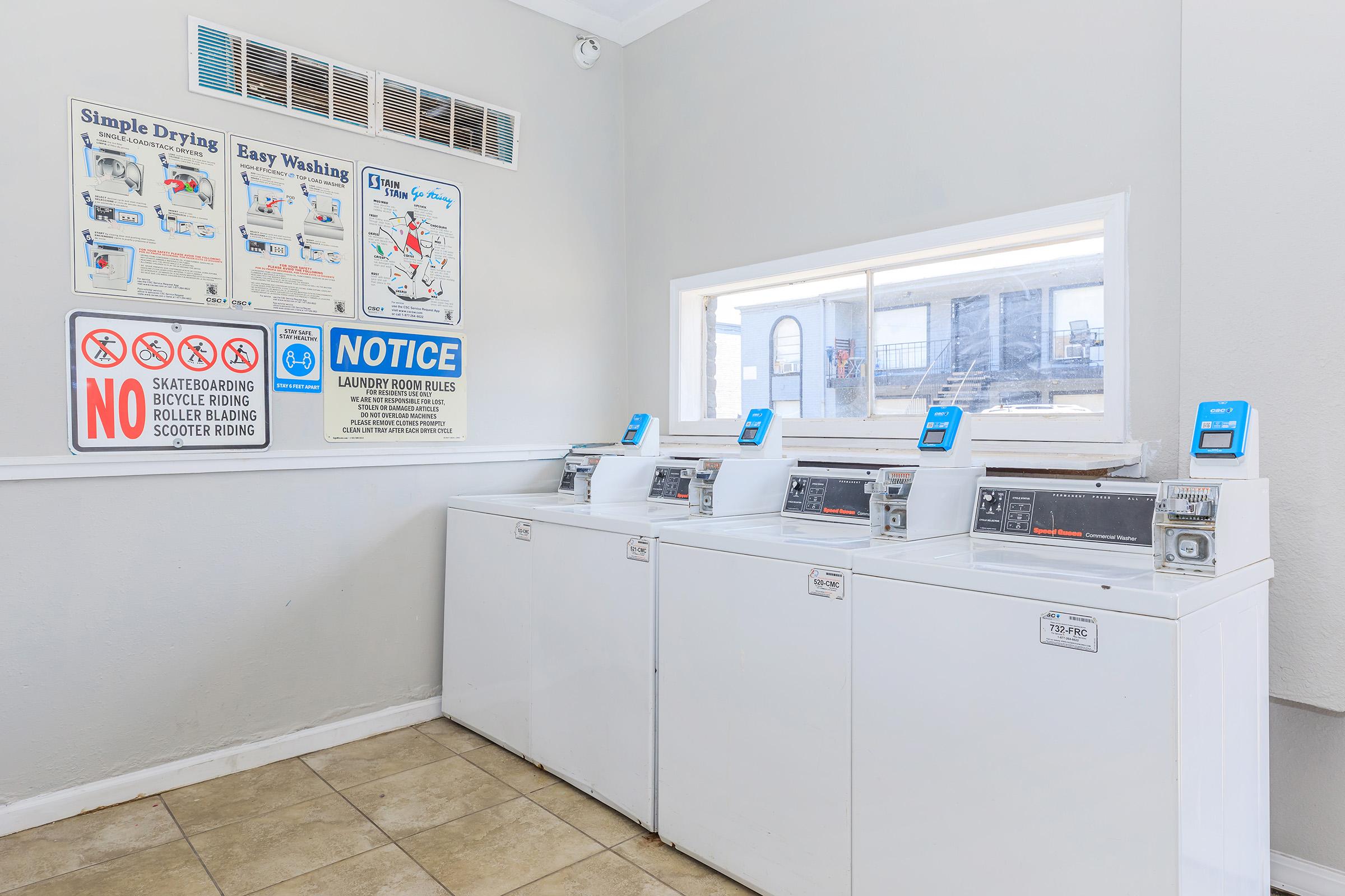 A bright laundromat featuring three row washers, each with a digital screen, against a light-colored wall. Nearby, signs provide washing instructions and laundry rules. A large window lets in natural light, highlighting the clean, tiled floor.