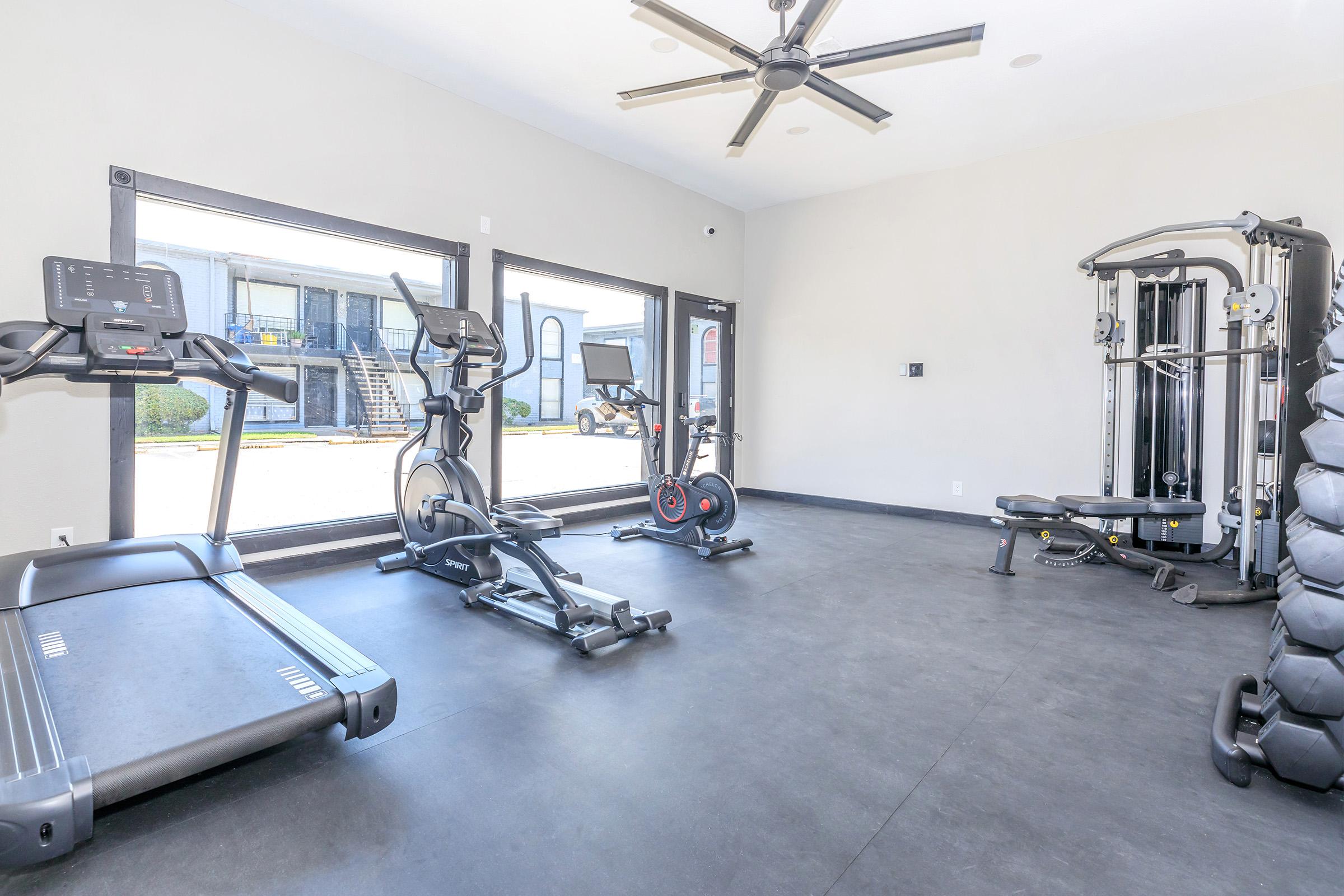 A modern gym with various exercise equipment, including treadmills, ellipticals, and a multi-functional weight machine. Large windows allow natural light to enter, providing a view of the outside area. The flooring is black, and the room is spacious and well-lit.