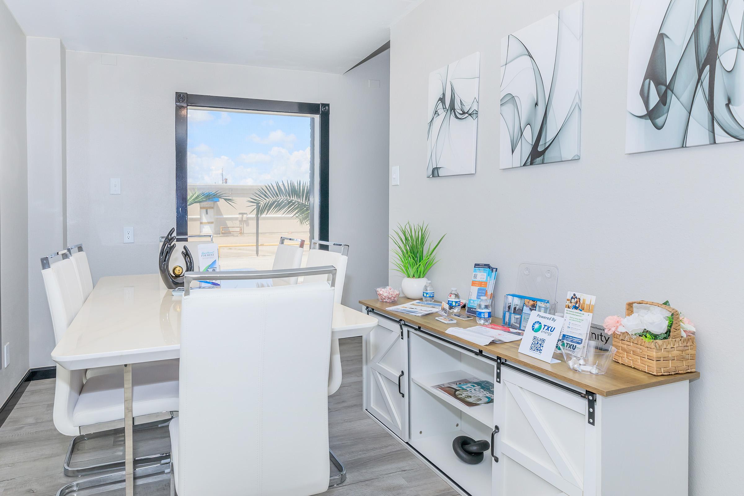 A modern dining area featuring a white table with six chairs, a large window letting in natural light, and abstract art on the walls. There’s a side console with decorative items, plants, and brochures, creating a welcoming and organized space.