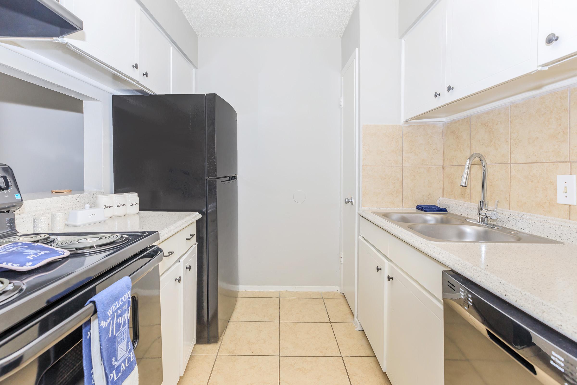 Modern kitchen featuring a black refrigerator, white cabinets, and beige tile floor. The countertop includes a sink, a stovetop, and a small appliance. A kitchen towel hangs from the oven. The decor is simple and functional, with ample natural light streaming in from an adjacent room.
