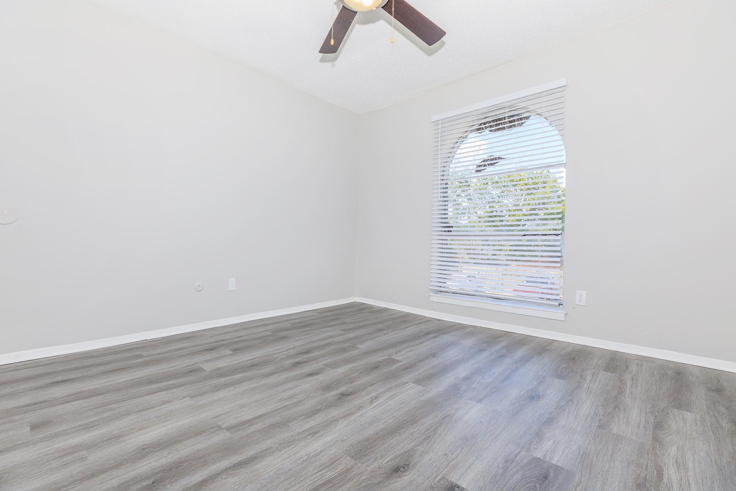 Empty room with light gray walls and a ceiling fan. The floor is covered with laminate wood-like flooring. A large arched window with white blinds allows natural light to enter the space, and there are no furnishings or decorations present.