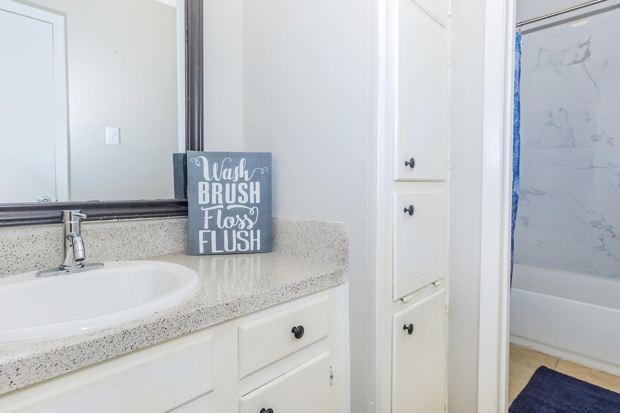 A bathroom interior featuring a white sink on a speckled countertop, a mirror above, and a decorative sign that reads "Wash, Brush, Floss, Flush." The space includes white cabinetry and a blue-tiled bathtub in the background, enhancing the clean and modern aesthetic.