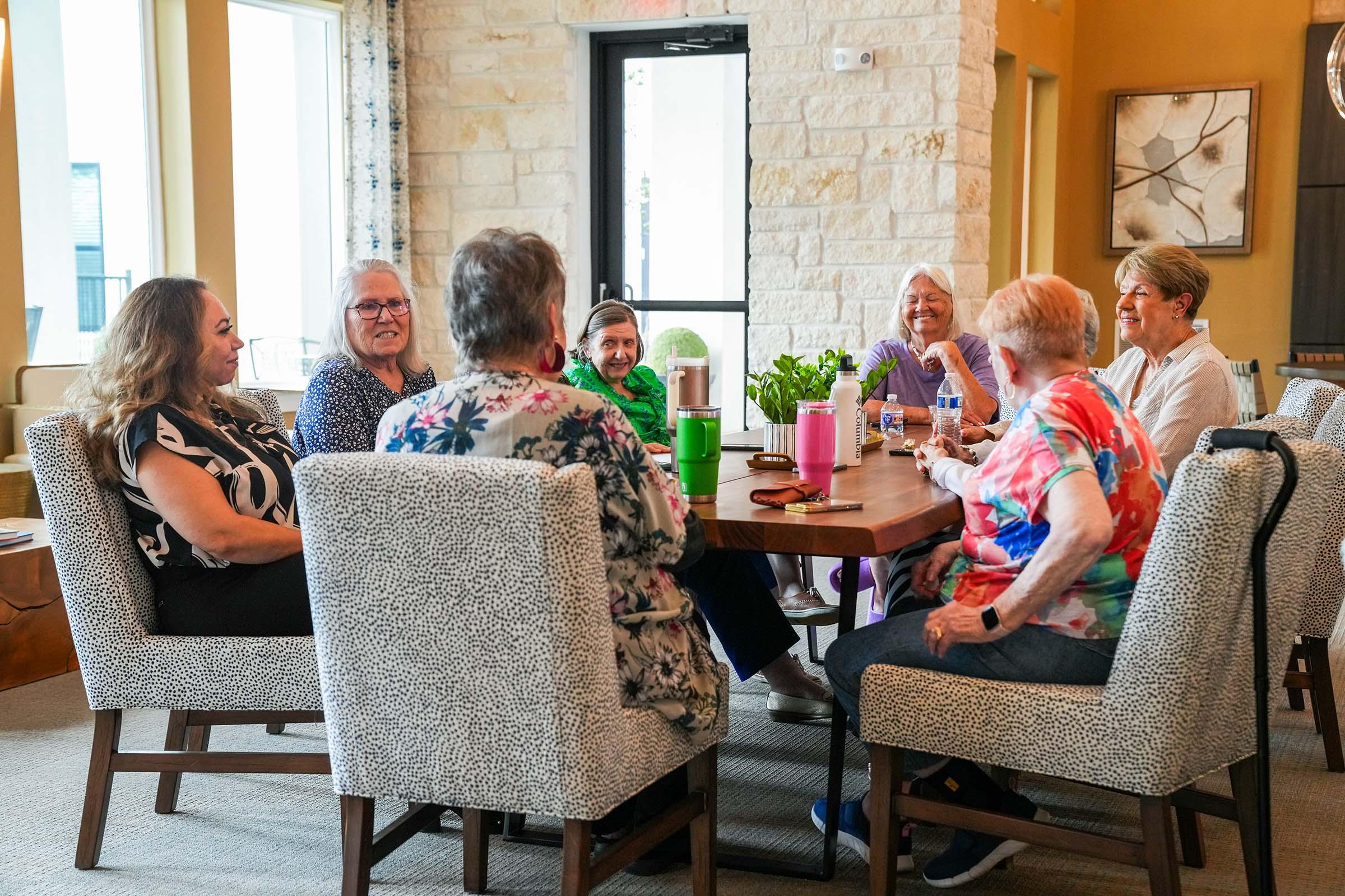 A group of eight women of varying ages sits around a large table in a bright, inviting room. They are engaged in conversation, smiling and sharing stories. The women are dressed in colorful clothing, and the table has drinks and a small plant centerpiece. Large windows let in natural light, enhancing the warm atmosphere.