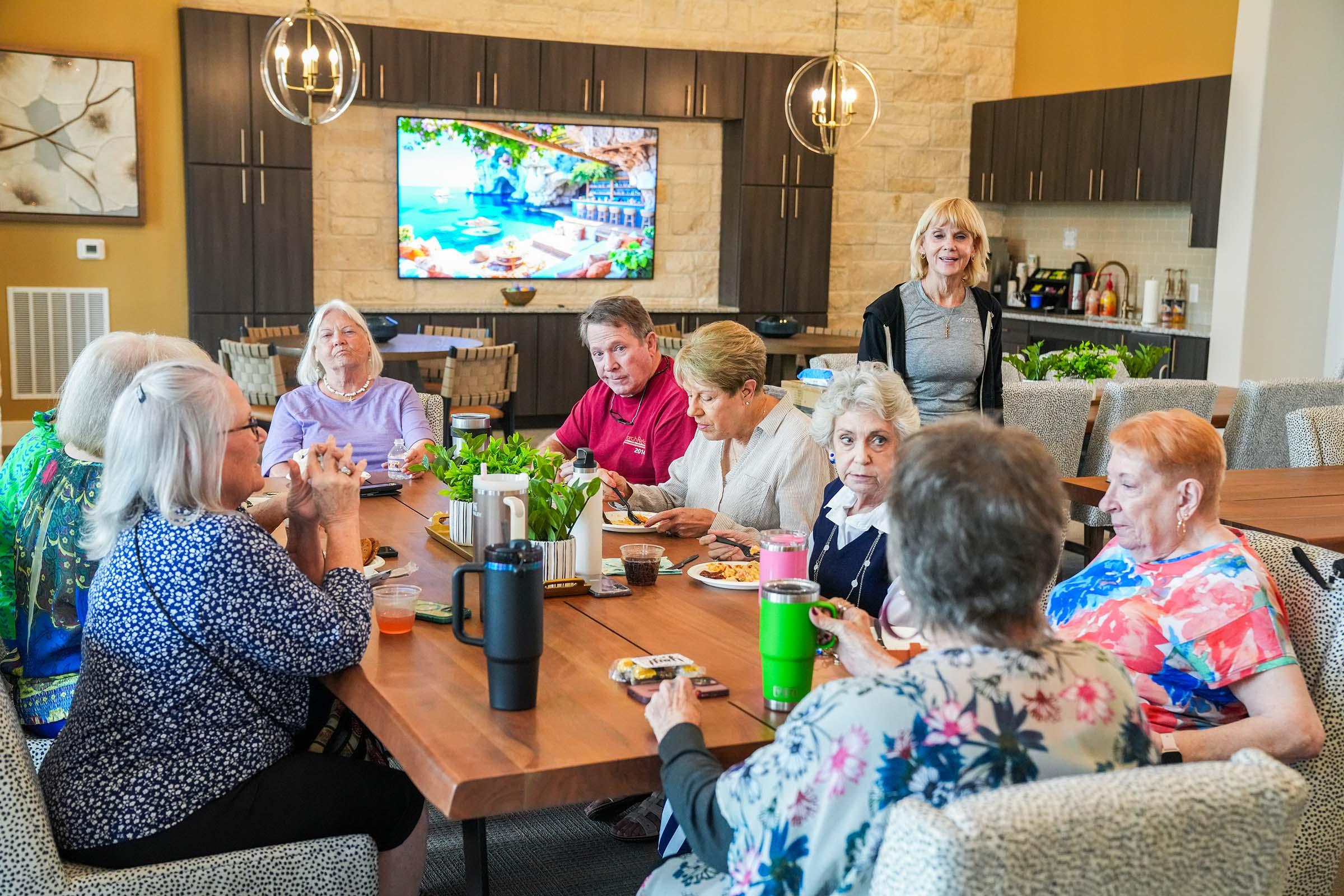 A group of seniors enjoying a meal together around a large dining table, with a variety of dishes and drinks. Some members are engaged in conversation, while others are eating. The background features a large screen displaying a scenic image. The setting has a warm, inviting atmosphere.