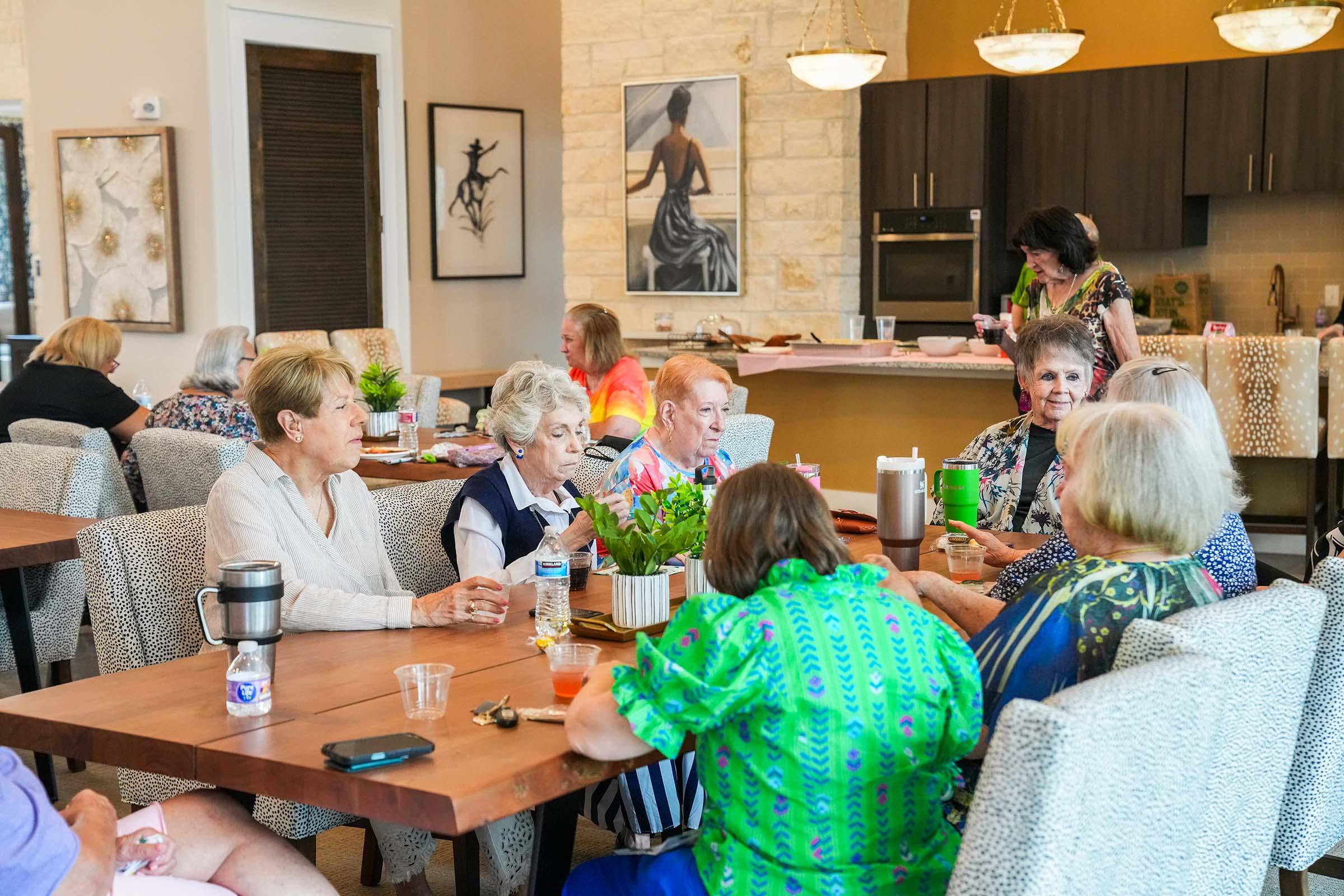 A group of seniors gathered around tables in a bright, modern community space. Some are chatting while others are enjoying refreshments. Decorative plants adorn the tables, and the background features a stylish kitchen and artwork on the walls. The atmosphere is lively and social.