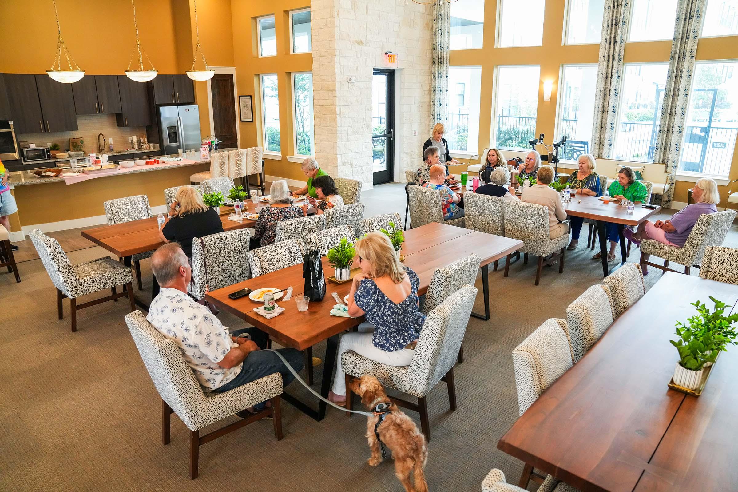 A spacious community dining area with various groups of seniors socializing at tables. Some are enjoying meals while others are engaged in conversation. A few plants decorate the tables, and a small dog on a leash is seen near one of the tables, adding to the lively atmosphere.