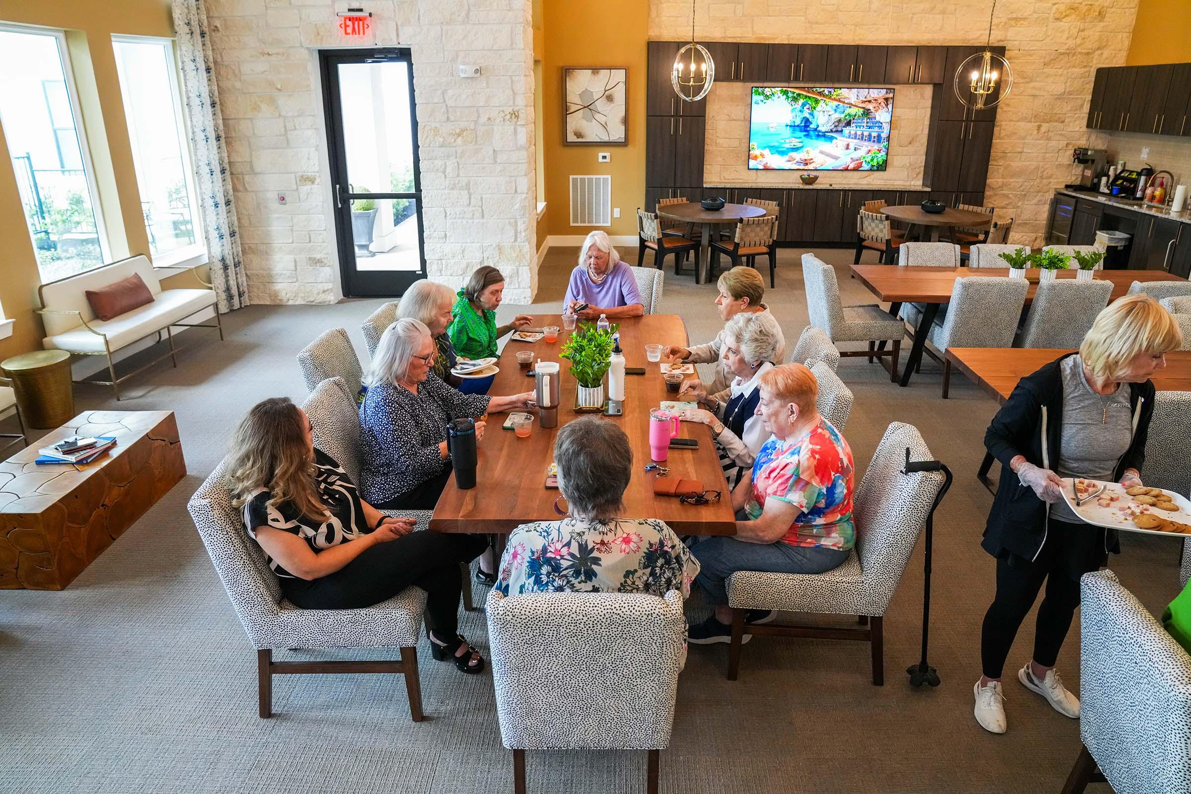 A group of older adults gathered around a large table in a well-lit dining area, enjoying a meal together. The room features large windows with natural light, modern decor, and a television displaying a colorful scene. Some individuals are engaged in conversation while others are eating.