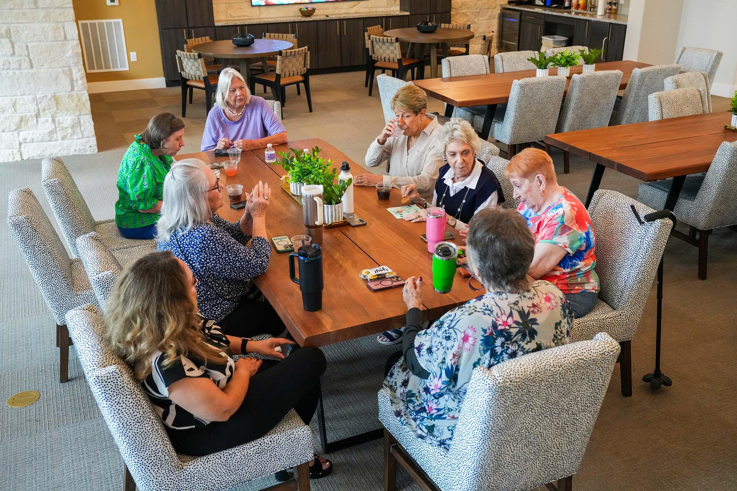 A group of eight elderly women sitting around a large wooden table in a communal space. They are engaged in conversation, enjoying refreshments, and wearing colorful clothing. The room features modern decor with light-colored walls, wooden furniture, and potted plants, creating a warm and inviting atmosphere.