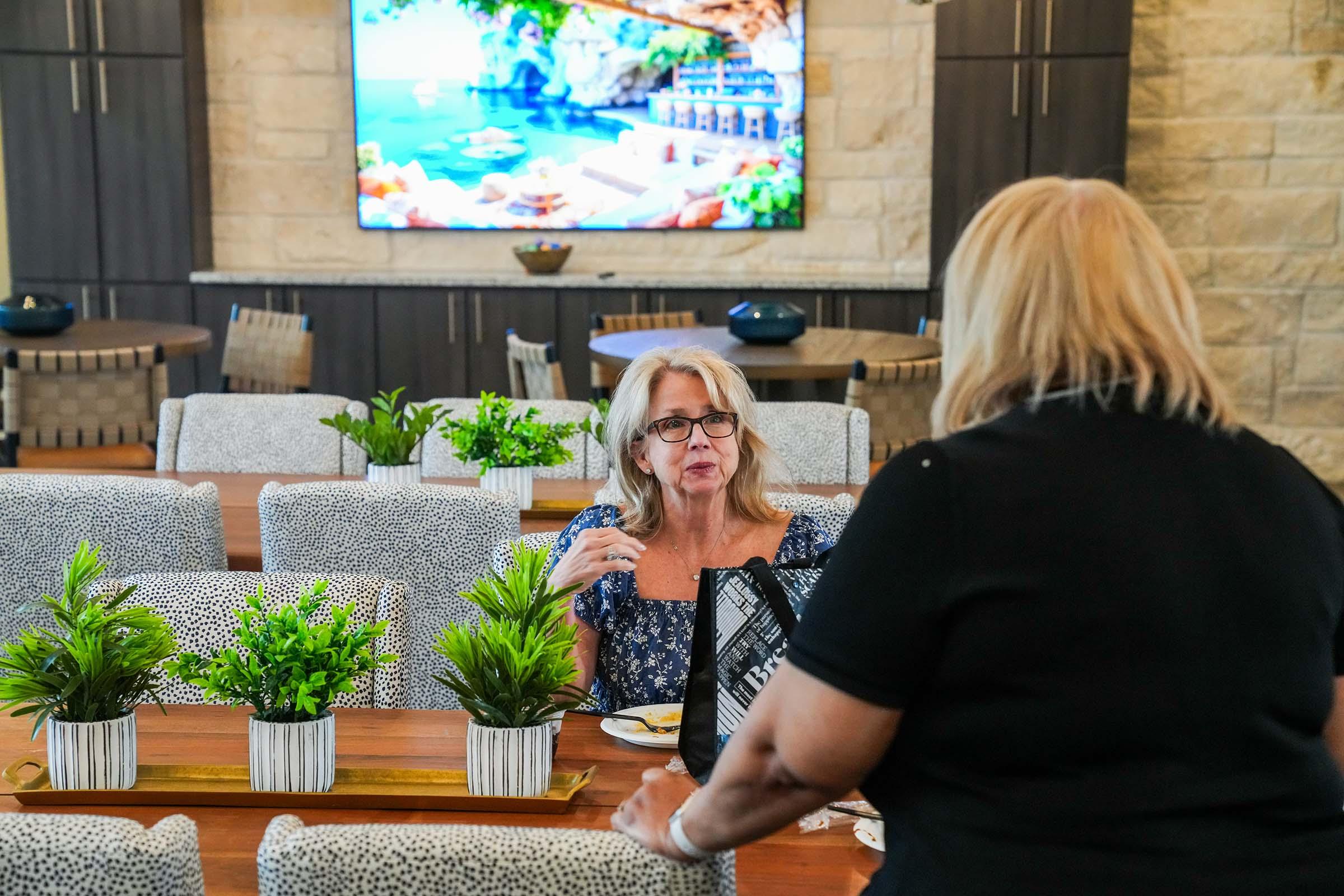 A woman with long, gray hair is sitting at a dining table, engaging in conversation with another woman. The background features a large screen displaying a scenic view of a beach, while small potted plants decorate the table. The setting appears cozy and contemporary.